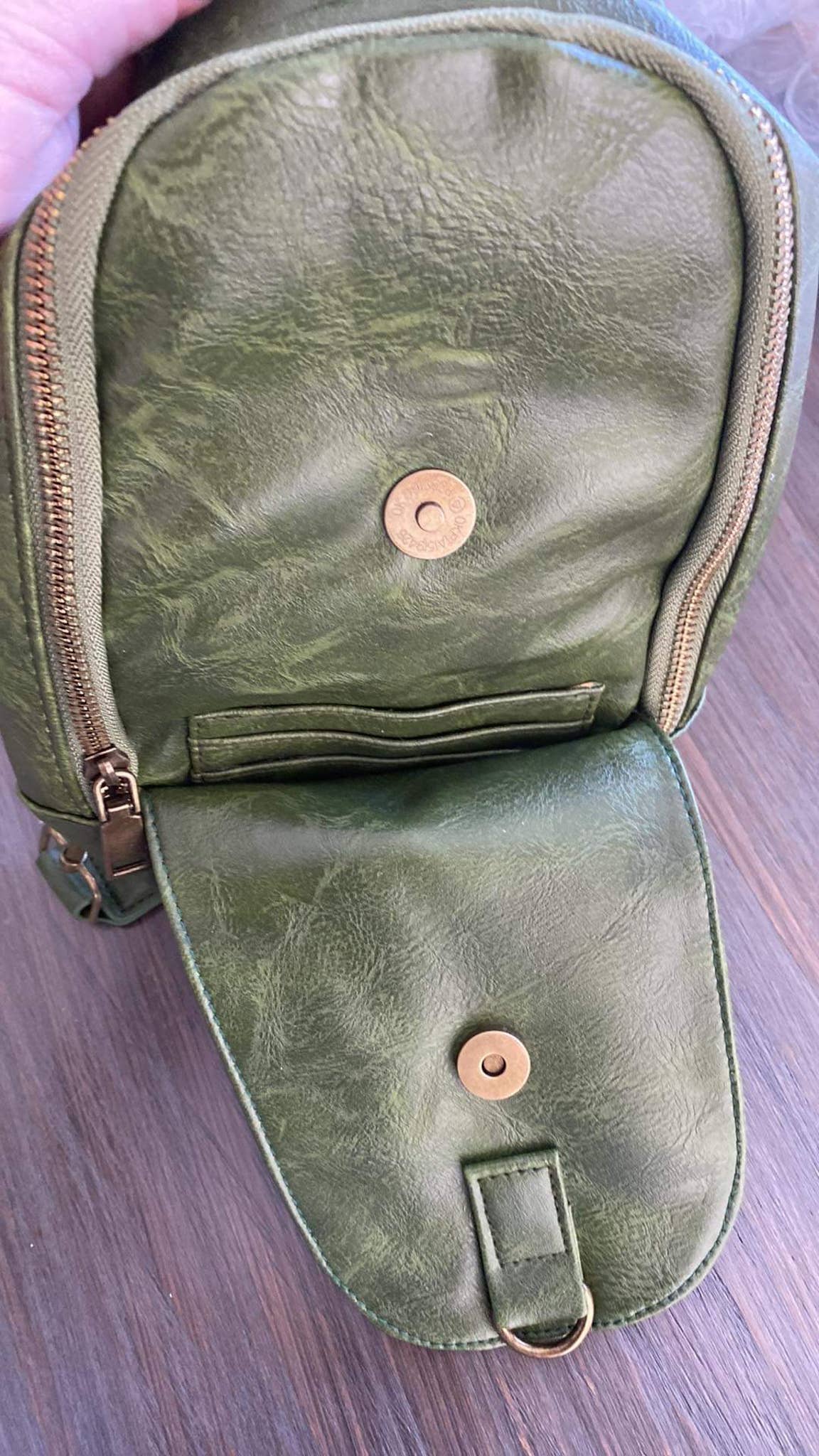 Alt text: A beige faux-leather sling bag with a zipper closure and a patterned strap featuring a tan and white zigzag diamond design. The bag is displayed on a dark wooden surface, showcasing its minimalist and stylish design.
