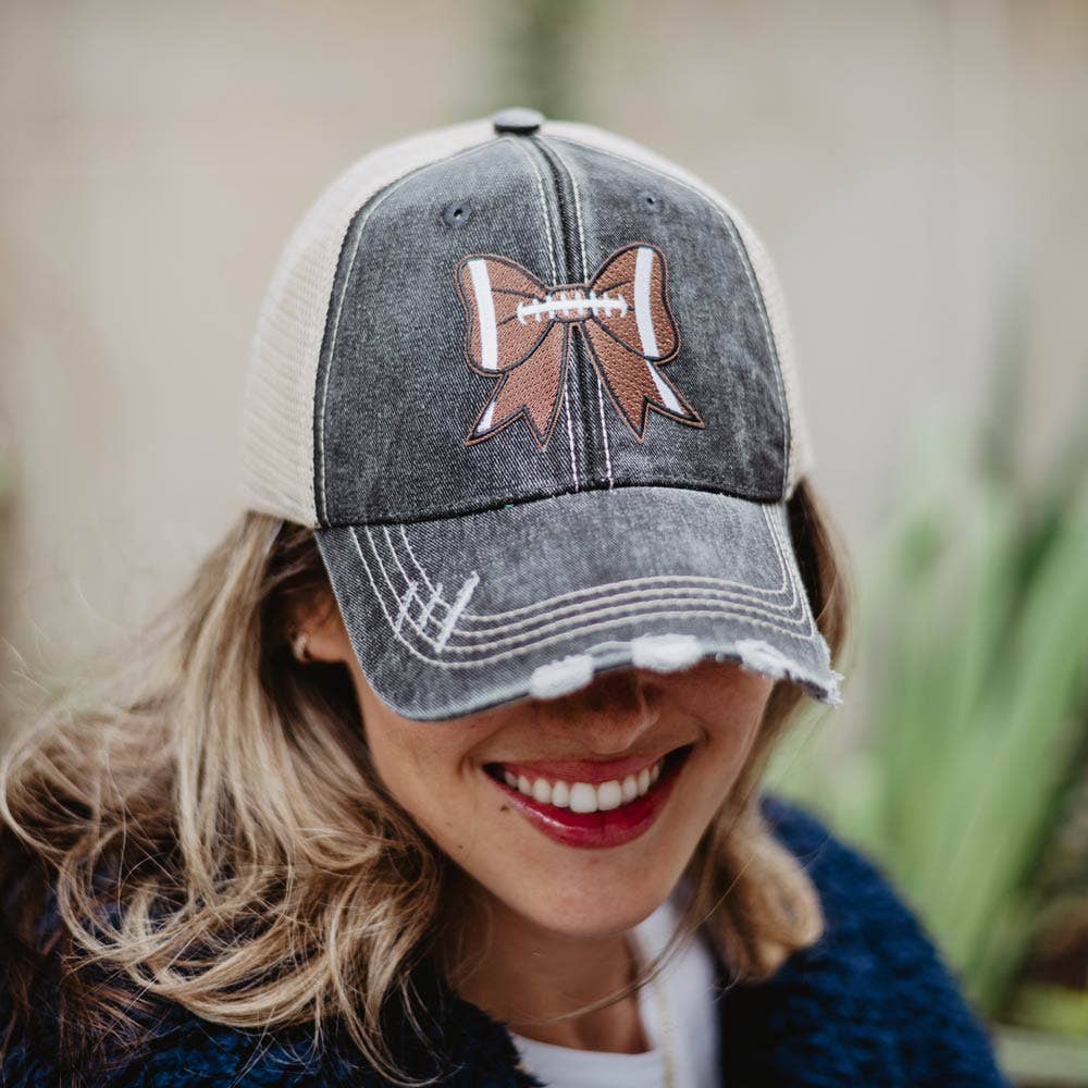 Distressed black and beige trucker hat featuring a football-themed bow patch on the front. Worn by a smiling woman, this stylish cap combines sporty and feminine elements, perfect for game days or casual outings.