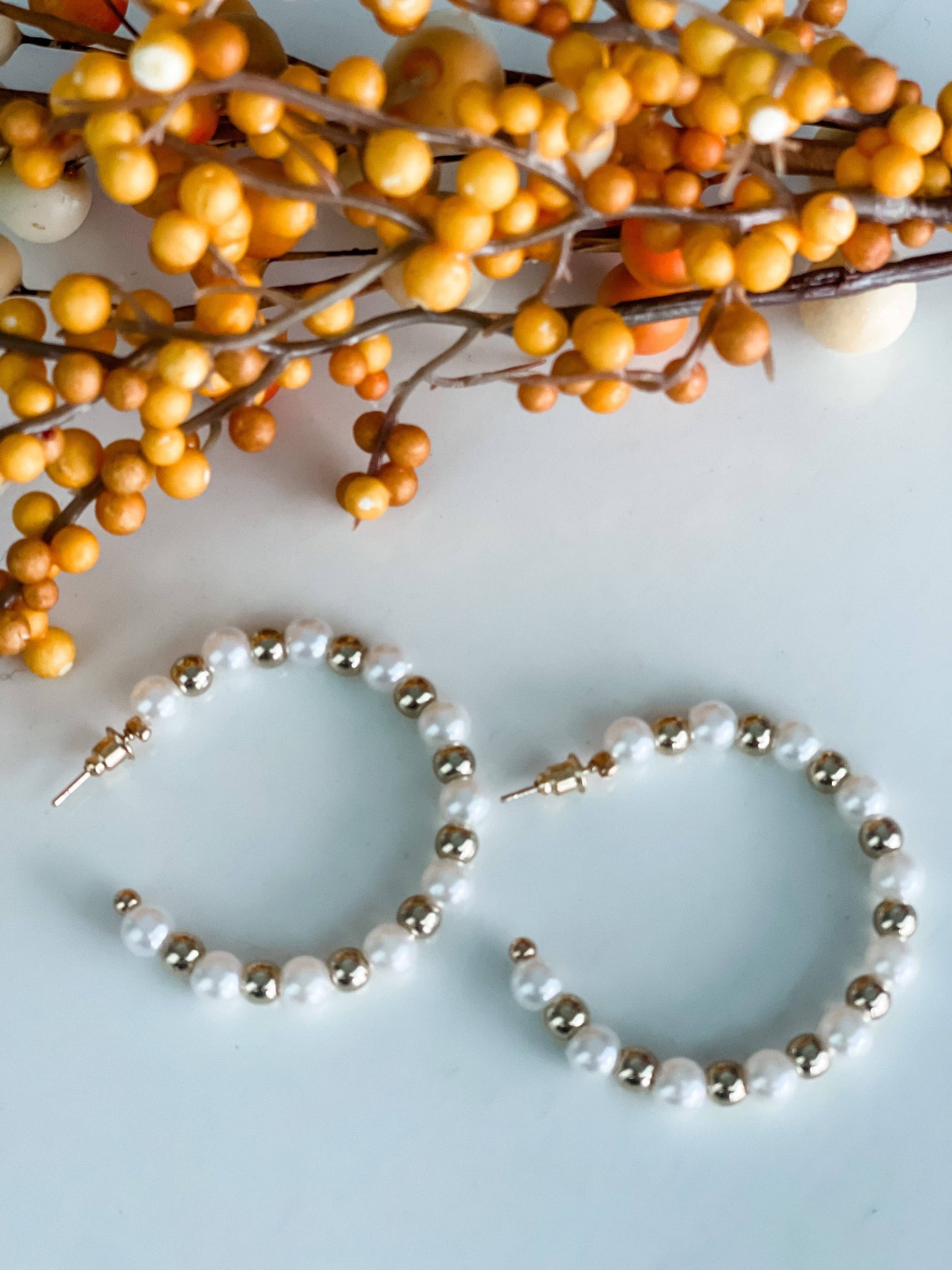 A pair of stylish hoop earrings featuring a mix of glossy white faux pearls and shiny gold beads, arranged in an alternating pattern. The earrings are displayed on a white surface with a decorative sprig of orange and yellow faux berries in the background, adding a warm, autumnal touch to the composition.