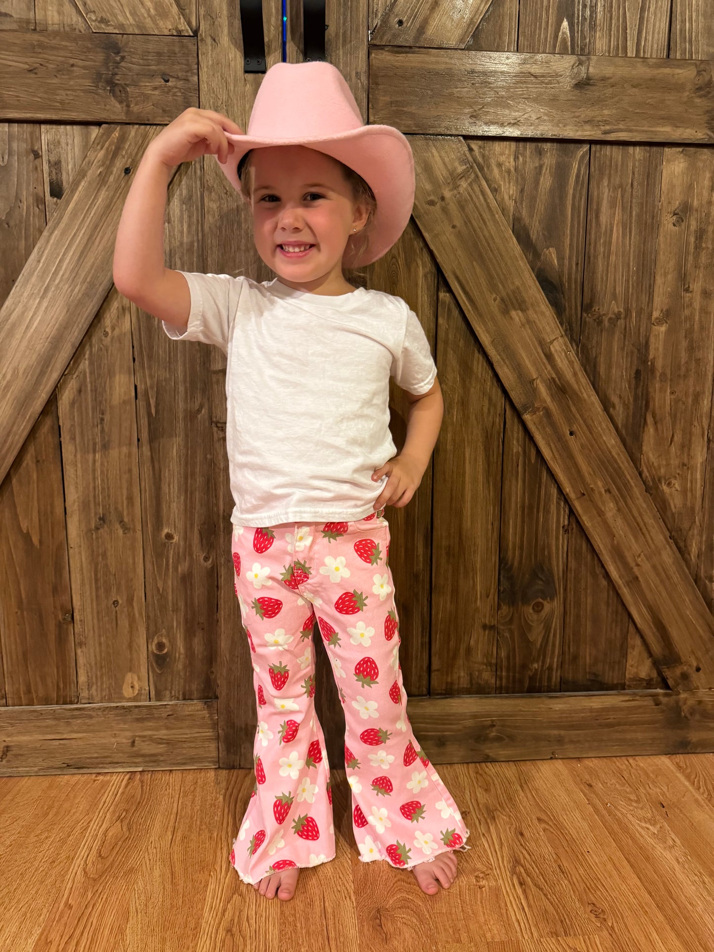 A smiling young girl wearing pink flared pants with a strawberry and daisy pattern, paired with a white top and a pink cowboy hat. She stands confidently in front of wooden barn-style doors, radiating charm and energy.