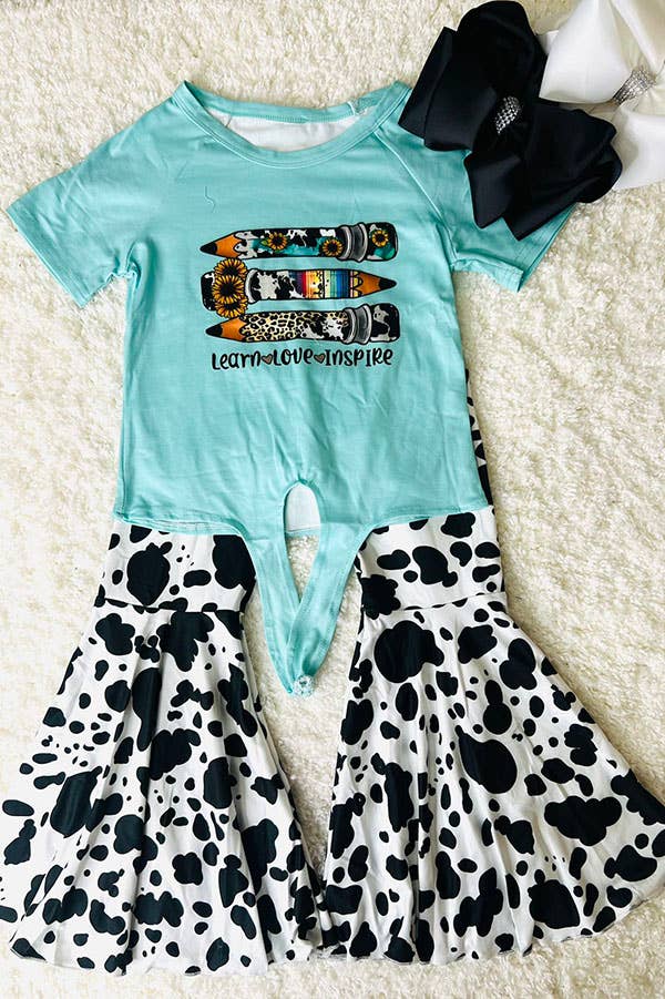 Trendy children's outfit featuring a turquoise top with a graphic design of decorative pencils and the phrase 'Learn Love Inspire' in playful black font. The top is paired with flared bell-bottom pants in a bold black-and-white cow print pattern. Complemented by oversized black and white hair bows with rhinestone accents, this ensemble is perfect for school or special occasions.