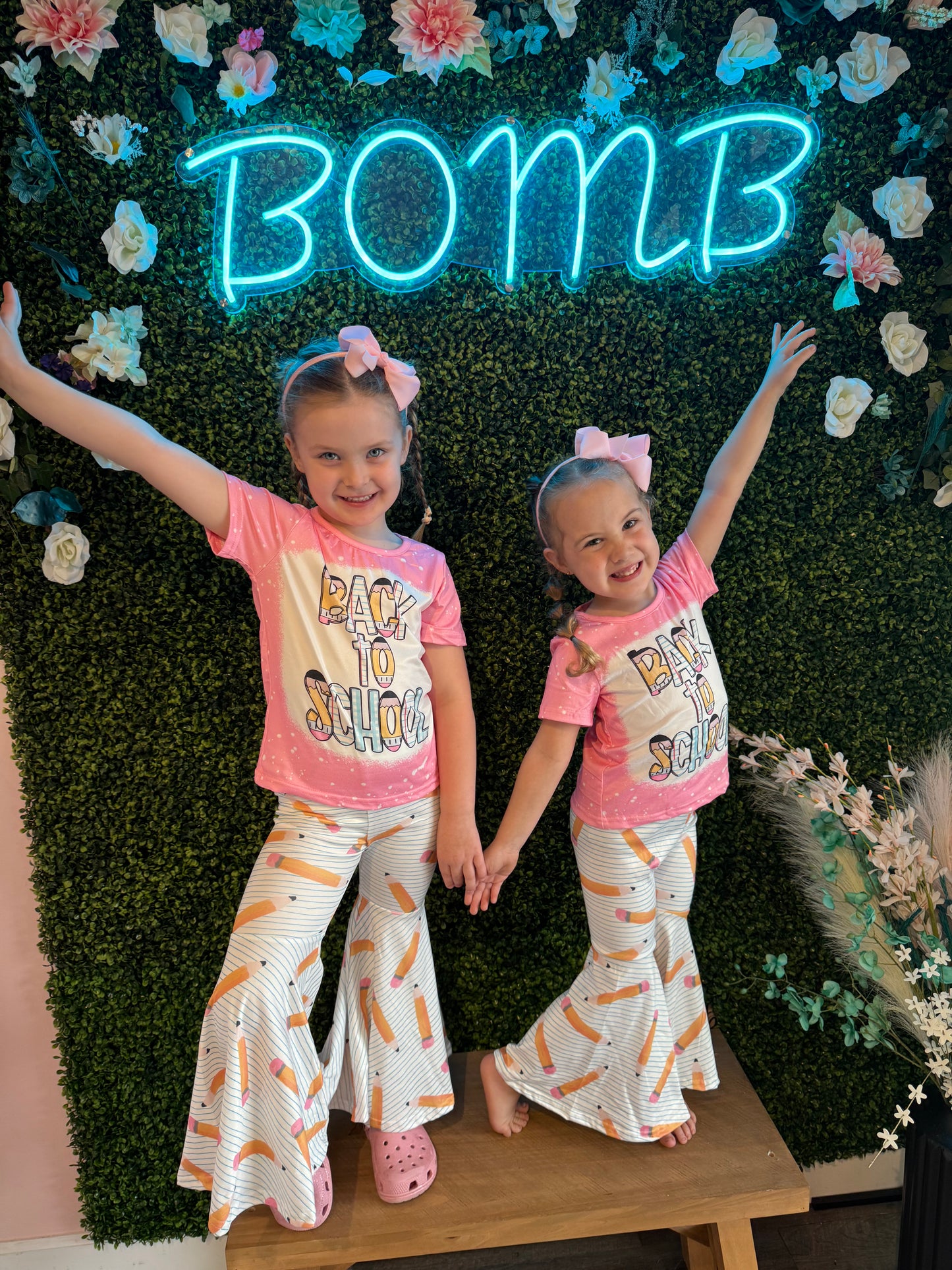 Two smiling young girls posing together in matching pink 'Back to School' T-shirts and white bell-bottom pants with a whimsical pencil pattern. They stand in front of a vibrant green faux foliage wall accented by flowers and a neon 'BOMB' sign, exuding a playful back-to-school vibe.