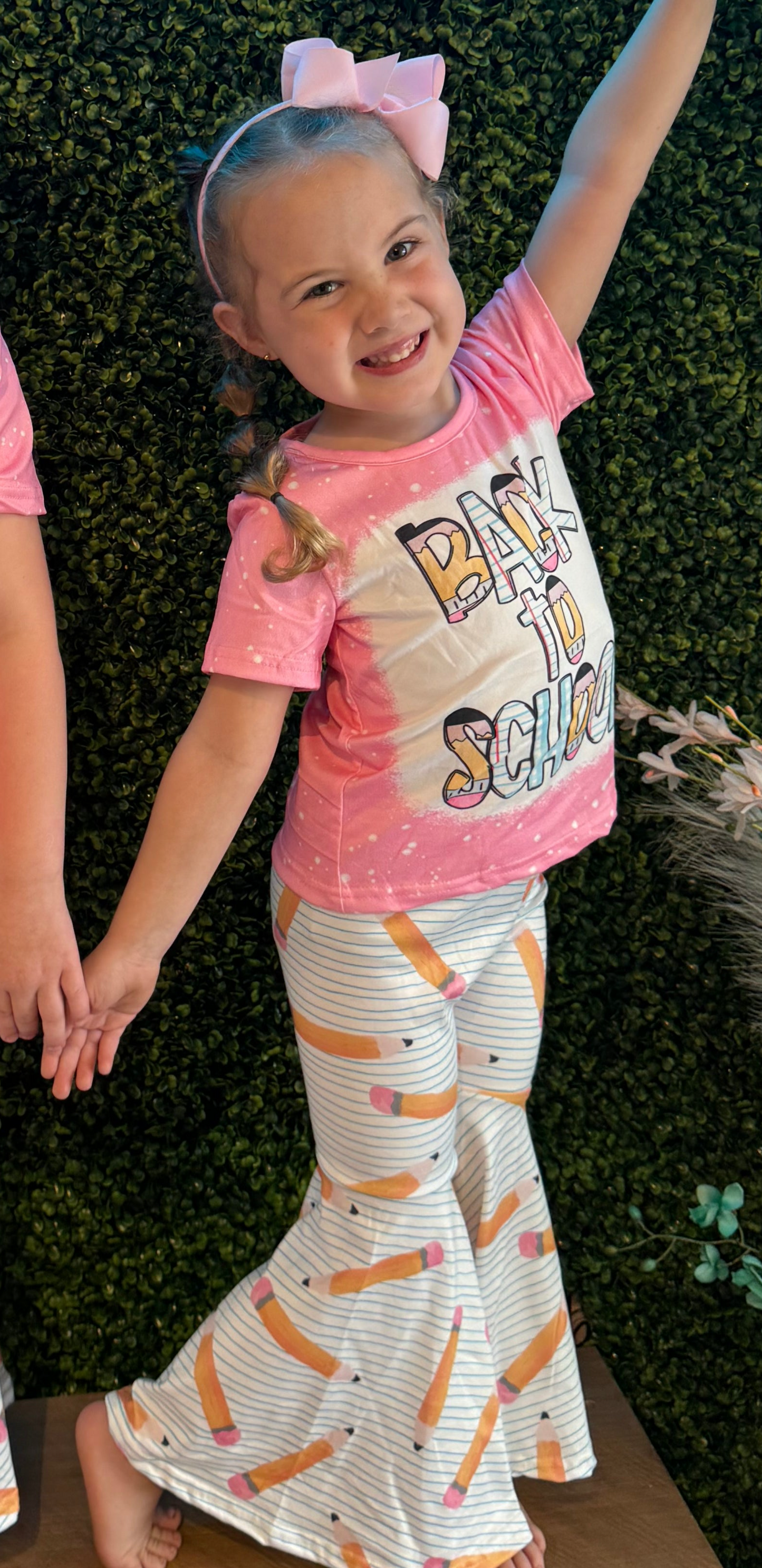 Smiling young girl posing in a pink tie-dye T-shirt with the phrase 'Back to School' in bold, colorful letters. She pairs it with white bell-bottom pants featuring a playful pencil pattern and a large pink hair bow. The green faux foliage wall behind her adds a vibrant and fun background.