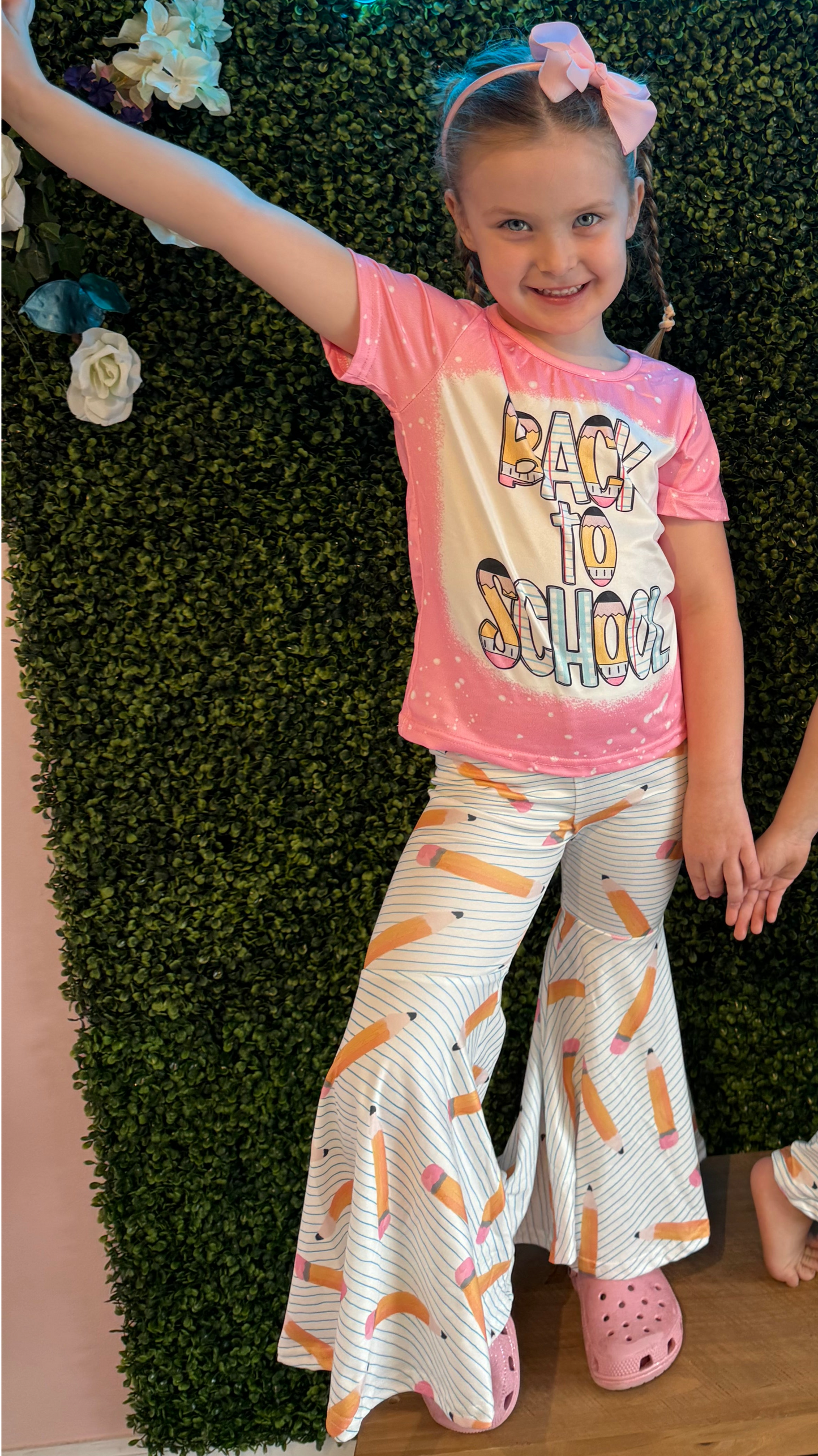 Young girl wearing a pink 'Back to School' T-shirt and white bell-bottom pants decorated with a pencil print. She strikes a cheerful pose, complemented by her pink bow and the lush green foliage wall adorned with white flowers in the background.