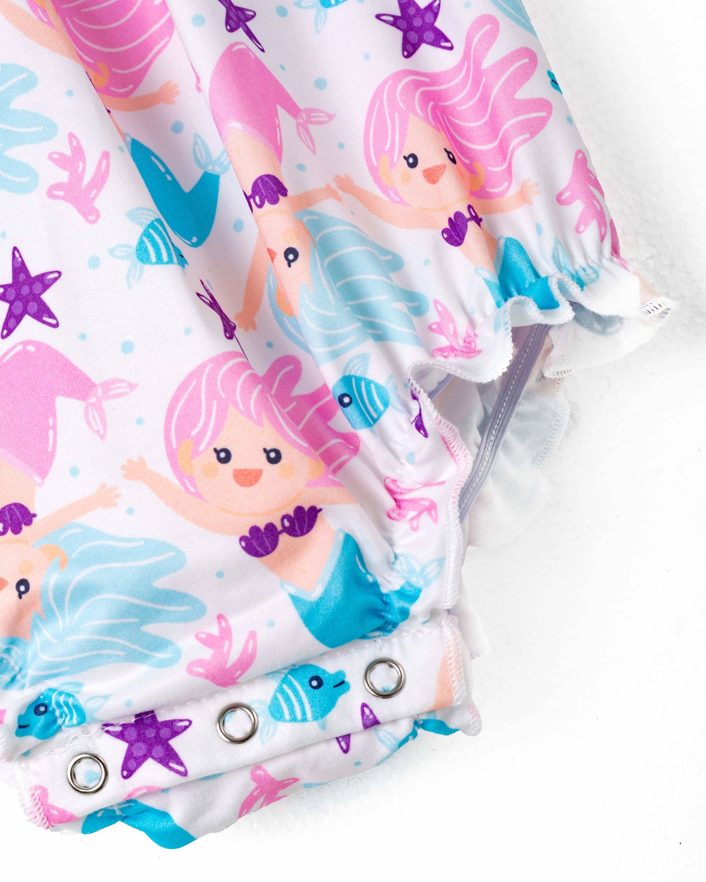 A colorful baby romper featuring a whimsical underwater mermaid theme. The fabric is adorned with pink and blue mermaids, starfish, fish, seashells, and coral motifs against a white background. The romper has bright blue straps tied into playful bows at the shoulders, adding a charming touch. The bottom has snap closures for easy diaper changes, and the gathered elastic at the legs ensures a comfortable fit. Displayed on a white hanger, the romper is perfect for summer or themed photoshoots.
