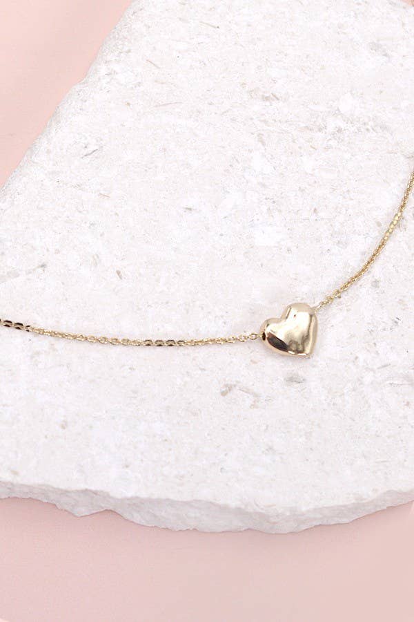 A close-up of a minimalist gold necklace featuring a delicate chain and a small polished heart-shaped pendant. The necklace is elegantly displayed on a textured white stone backdrop, emphasizing its simplicity and timeless charm, perfect for everyday wear or layering.