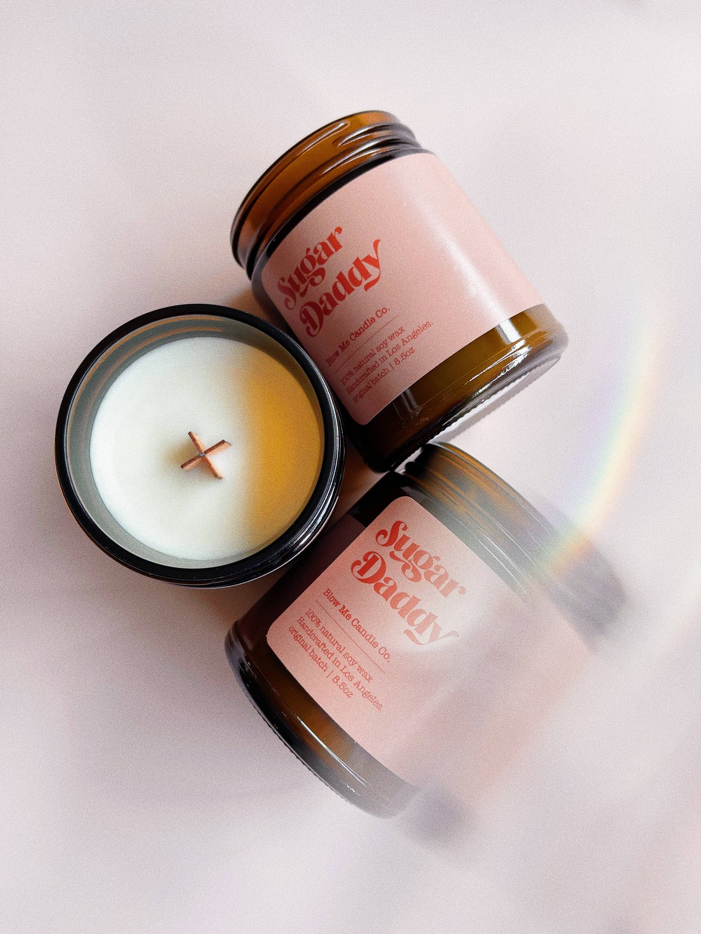 Three amber jar candles labeled "Sugar Daddy" by Blow Me Candle Co., with soft pink labels. One candle is open, revealing a wooden wick inside, while the others are displayed on their sides against a light background with a subtle rainbow reflection.