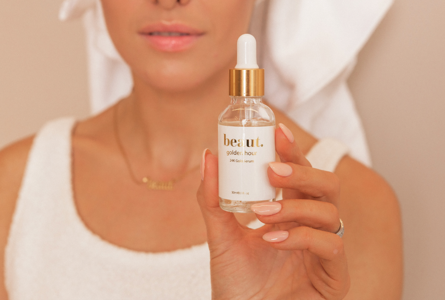 Model holding Beaut. Golden Hour 24K Gold Collagen Serum, showcasing its elegant glass bottle with visible gold flakes and a gold cap. The scene emphasizes natural beauty and self-care, with the model wearing a white towel and minimal makeup for a fresh, clean look.