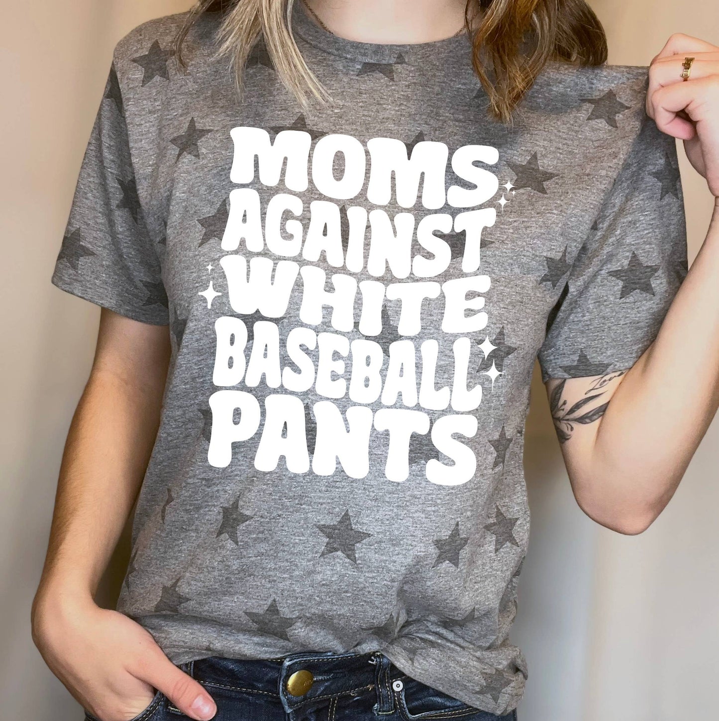 A gray t-shirt with black star patterns featuring bold white text that says "MOMS AGAINST WHITE BASEBALL PANTS." The shirt is styled casually with blue jeans, ideal for baseball moms.