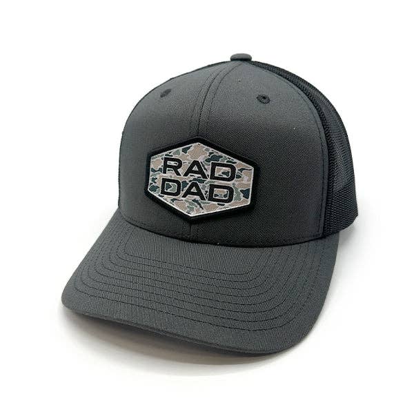 A dark gray trucker-style cap with a structured front and black mesh back. The front features a hexagonal patch with the words "RAD DAD" in bold, uppercase letters, set against a camouflage background. The curved brim and minimalist design emphasize a casual, modern style.