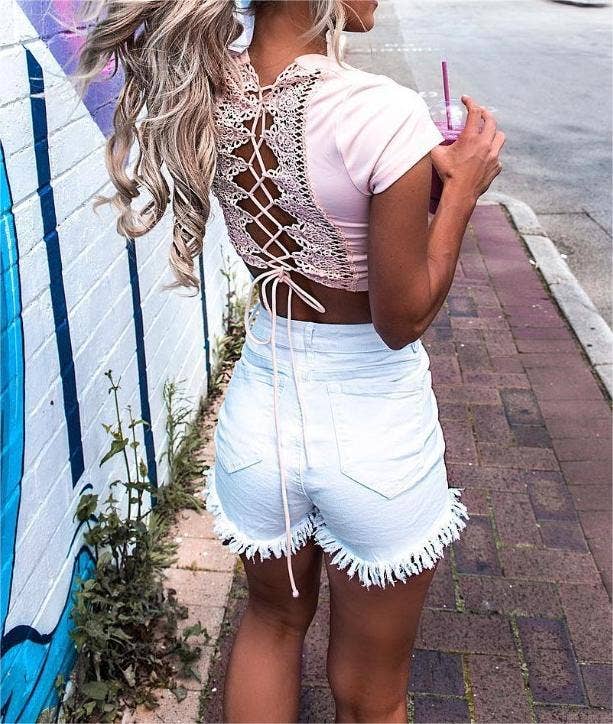 A model styled in high-waisted white frayed denim shorts paired with a pink crop top featuring lace-up back detailing, standing against an urban graffiti wall.
