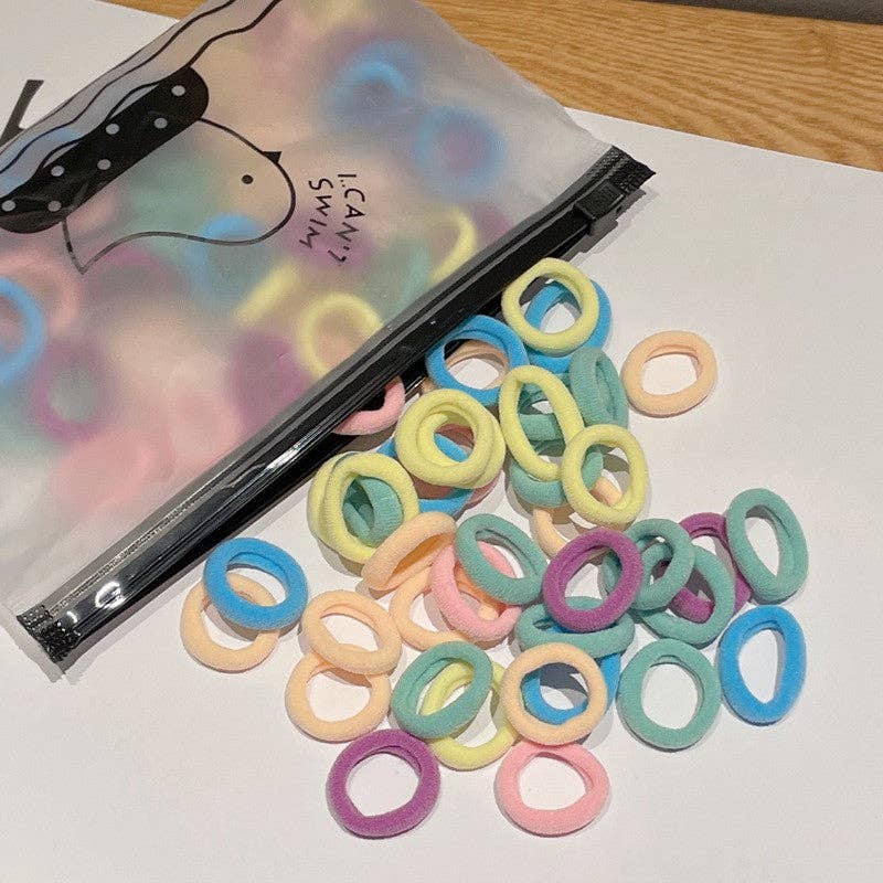 A small plastic zippered bag labeled "I Can Swim" is open, spilling out a colorful assortment of hair ties onto a white surface. The hair ties come in soft pastel shades, including pink, purple, yellow, green, and blue, creating a vibrant and playful display.