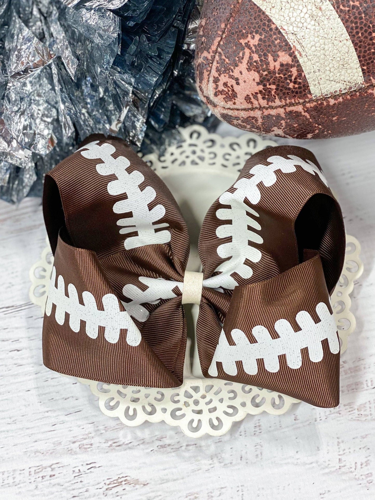Large brown grosgrain hair bow designed with white football lace detailing, perfect for game-day or sports-themed events. The bow is styled on a decorative background and shown in use atop a child's hairstyle, complementing a sporty outfit. Ideal for football fans and festive occasions.