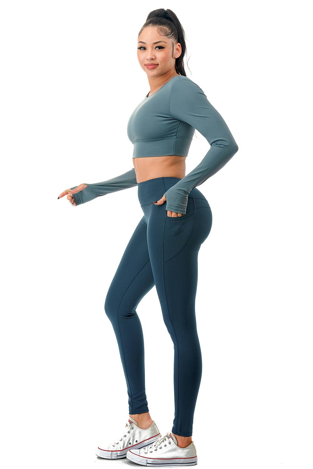 A model wearing a stylish teal two-piece activewear set consisting of a long-sleeve cropped top and high-waisted leggings. The top features a fitted design with thumbholes and a crisscross strap detail at the open back, providing both support and style. The leggings have a smooth, high-rise waistband with a seamless look, complementing the fitted silhouette. The model completes the outfit with white sneakers and a high ponytail, showcasing a sporty and chic appearance. Perfect for workouts, yoga,