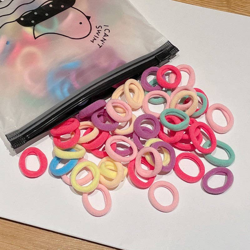 A small plastic zippered bag labeled "I Can Swim" is open, spilling out a colorful assortment of hair ties onto a white surface. The hair ties come in soft pastel shades, including pink, purple, yellow, green, and blue, creating a vibrant and playful display.