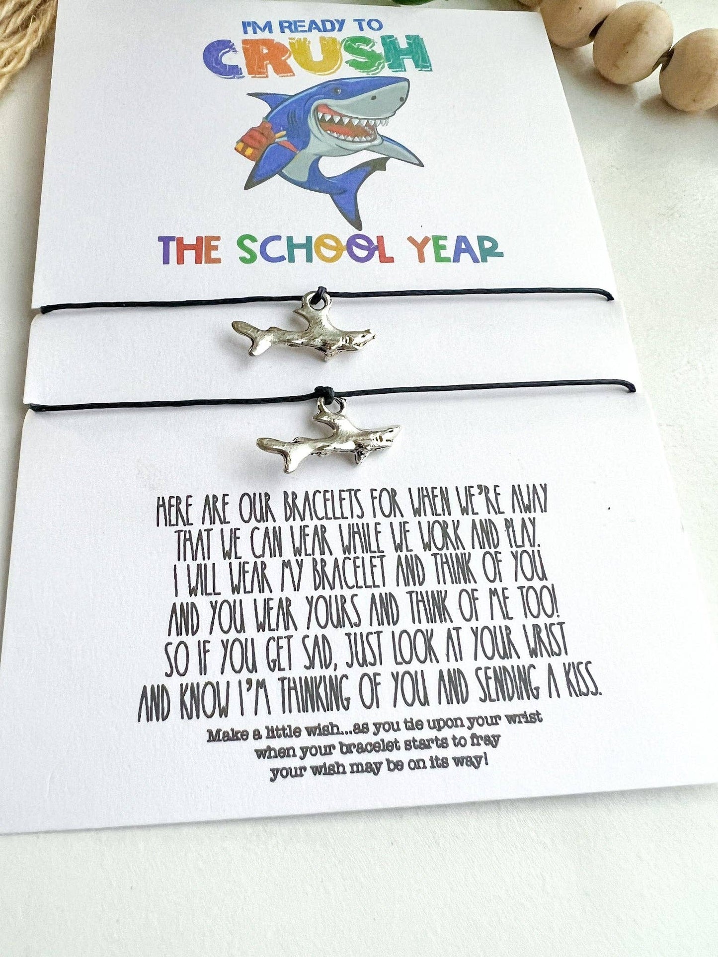 Close-up image of a back-to-school gift set featuring two matching shark charm bracelets on black cords. The bracelets are displayed on a white card with colorful text that reads "I'M READY TO CRUSH THE SCHOOL YEAR," accompanied by a playful illustration of a smiling shark wearing a backpack. Below the bracelets, a heartfelt poem encourages the wearer to think of their loved ones while apart, with a message of support and encouragement.