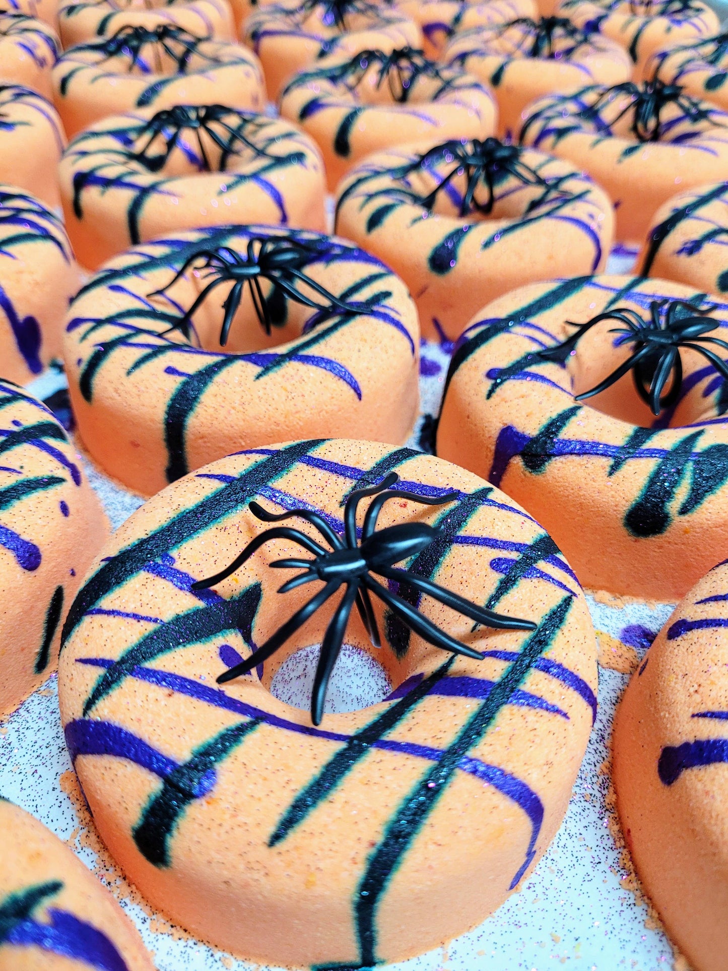 A collection of Halloween-themed orange bath bombs shaped like donuts, decorated with black plastic spiders and streaks of black and purple glitter. The vivid colors and creepy accents make them festive and perfect for seasonal celebrations.