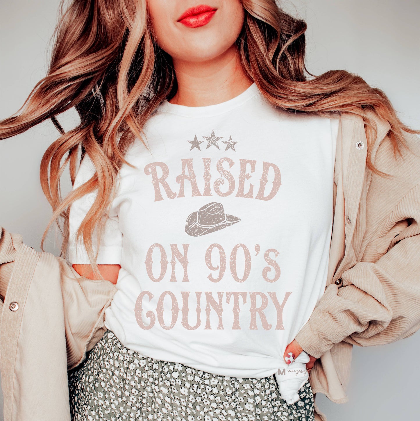 A white t-shirt with the phrase "RAISED ON 90'S COUNTRY" printed in a distressed, vintage-style font. The design includes a cowboy hat graphic below three small stars, enhancing its country-themed aesthetic. The model pairs the shirt with a floral-patterned skirt and a beige corduroy jacket, adding a casual, feminine touch. Loose waves and bold red lipstick complete the nostalgic, country-inspired look.