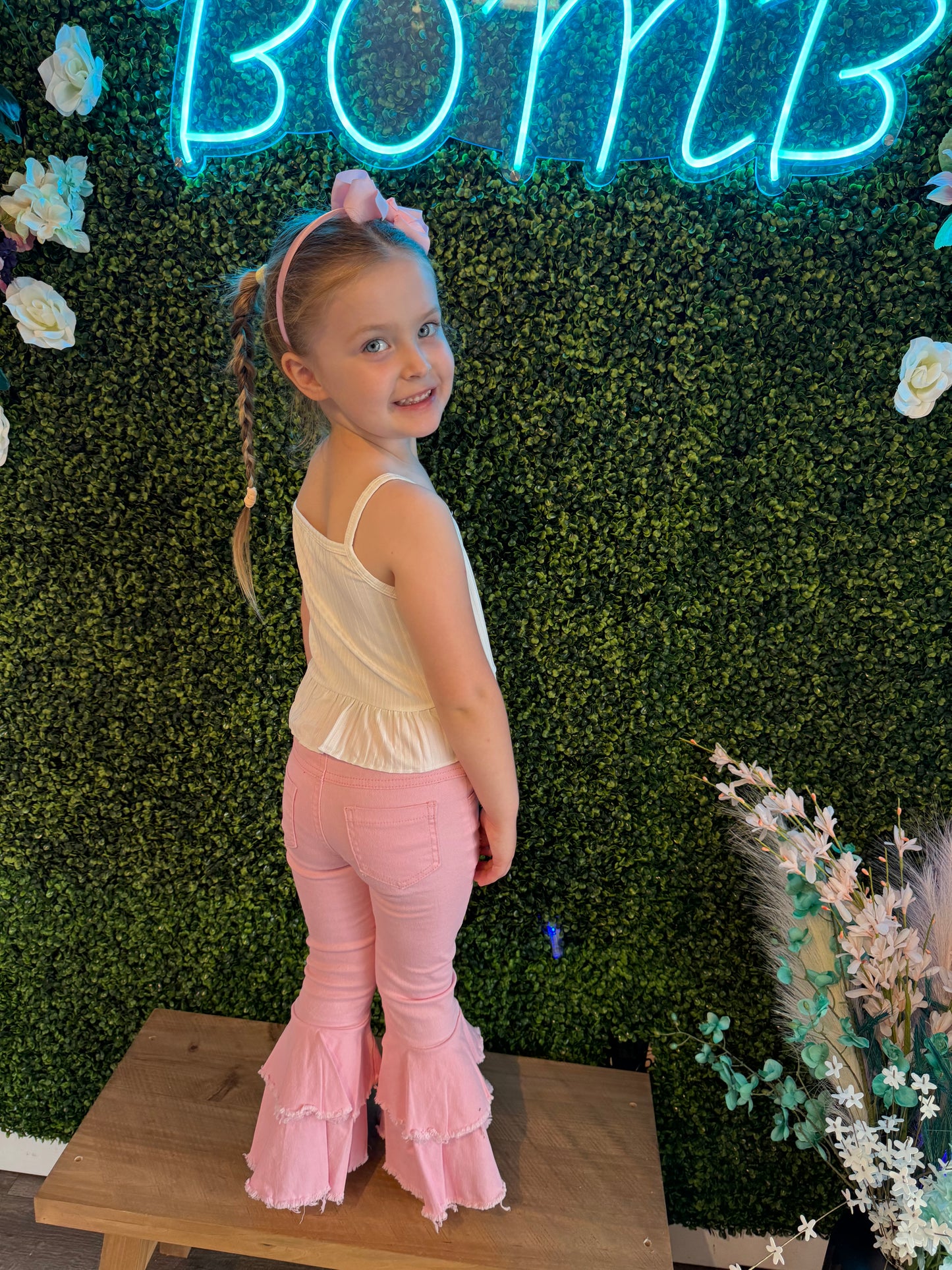 girl strikes a playful pose indoors, while standing barefoot on a wooden platform. She wears the white sleeveless top and pink bell-bottom pants and has her hair accessorized with a pink bow. She is framed by a decorative green faux foliage wall adorned with flowers, with a bright blue neon sign reading "BOMB" glowing above her. A floral arrangement in a black vase adds an elegant touch to the scene.