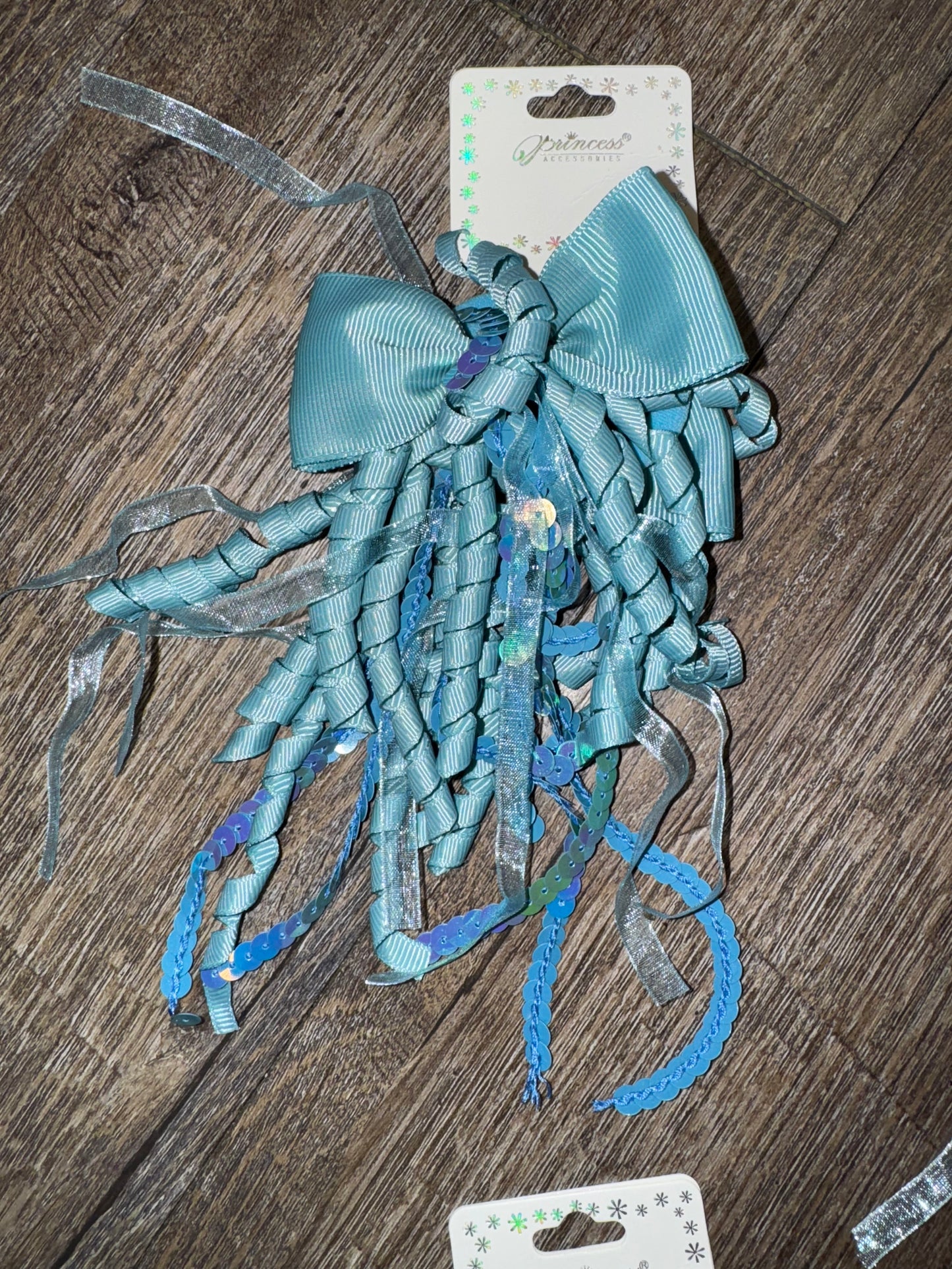 A set of light blue hair bows with cascading curly ribbons and sparkly sequin details, displayed on branded packaging labeled "Princess Accessories." The bows are placed on a wooden surface, showcasing their whimsical and elegant design.