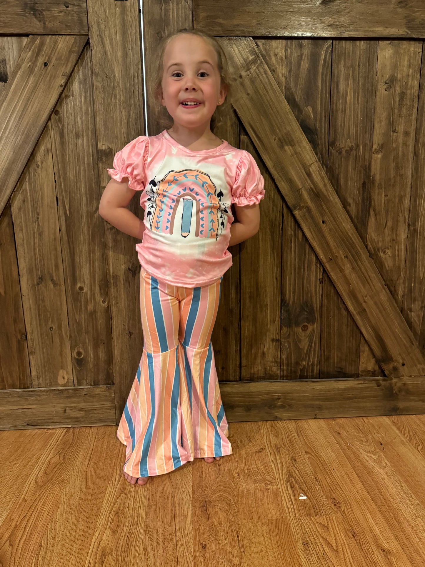 Smiling young girl wearing a pink puff-sleeve T-shirt featuring a colorful boho rainbow design with feather accents. She pairs it with flared bell-bottom pants in pastel pink, orange, and blue vertical stripes. The rustic wooden barn door background and warm wood flooring add a charming, country-inspired touch.