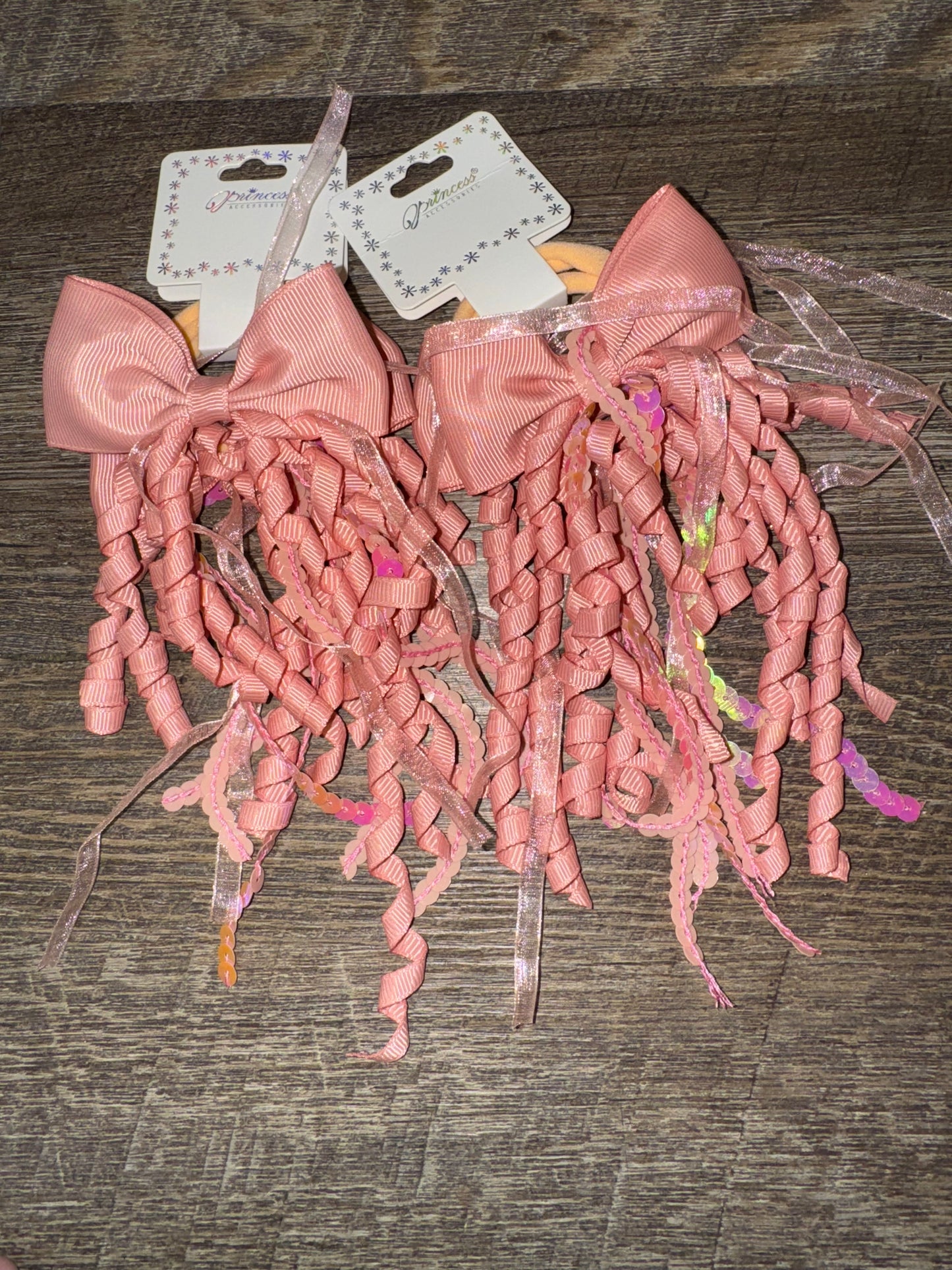 A set of light pink hair bows with cascading curly ribbons and sparkly sequin details, displayed on branded packaging labeled "Princess Accessories." The bows are placed on a wooden surface, showcasing their whimsical and elegant design.