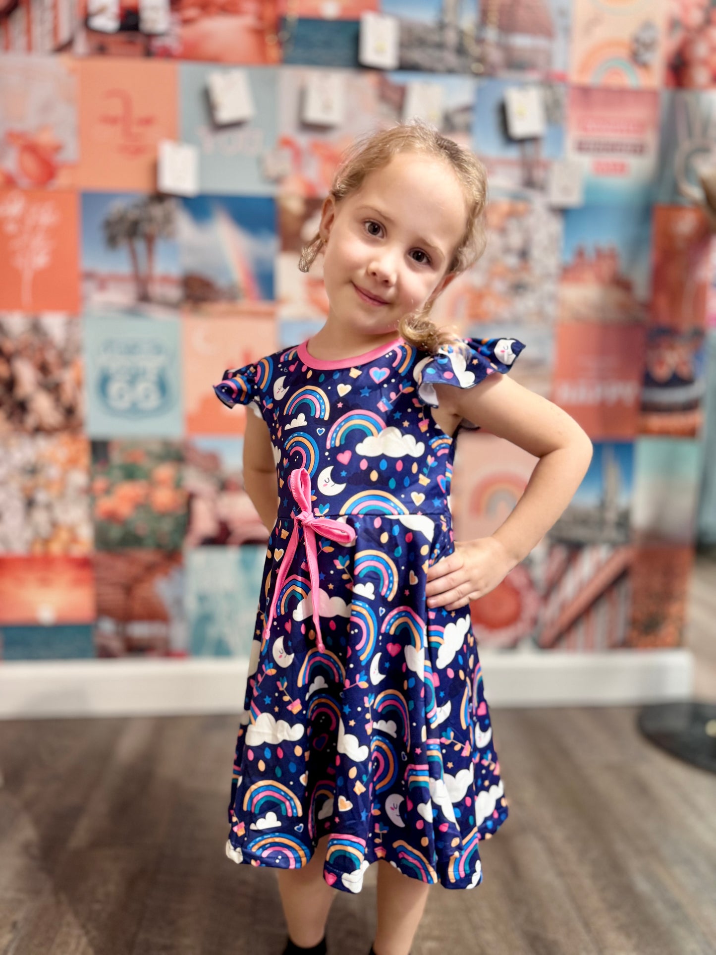 Smiling young girl in a navy dress with rainbow and cloud patterns, posing confidently in front of a vibrant backdrop.