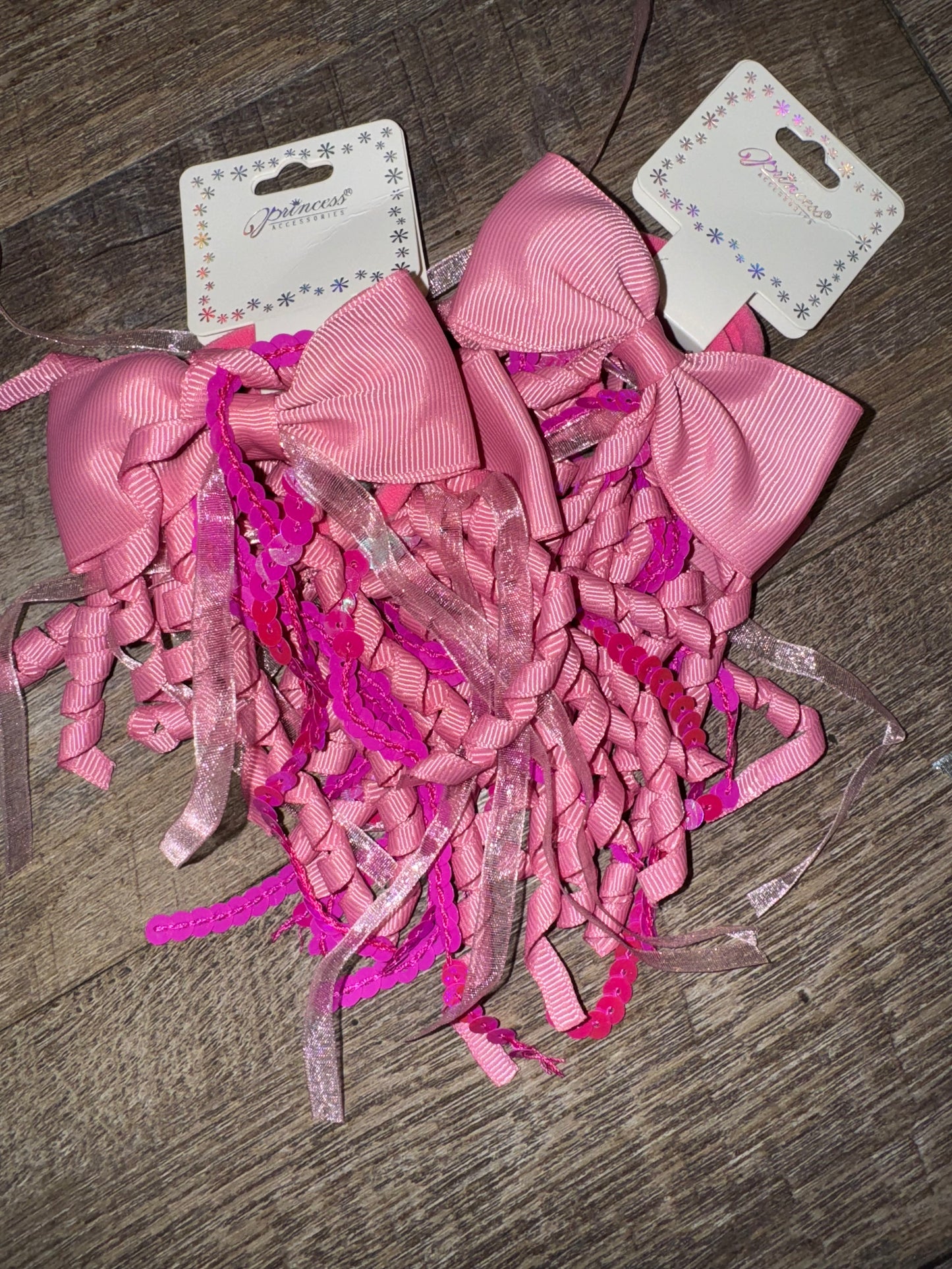 A set of light purple hair bows with cascading curly ribbons and sparkly sequin details, displayed on branded packaging labeled "Princess Accessories." The bows are placed on a wooden surface, showcasing their whimsical and elegant design.