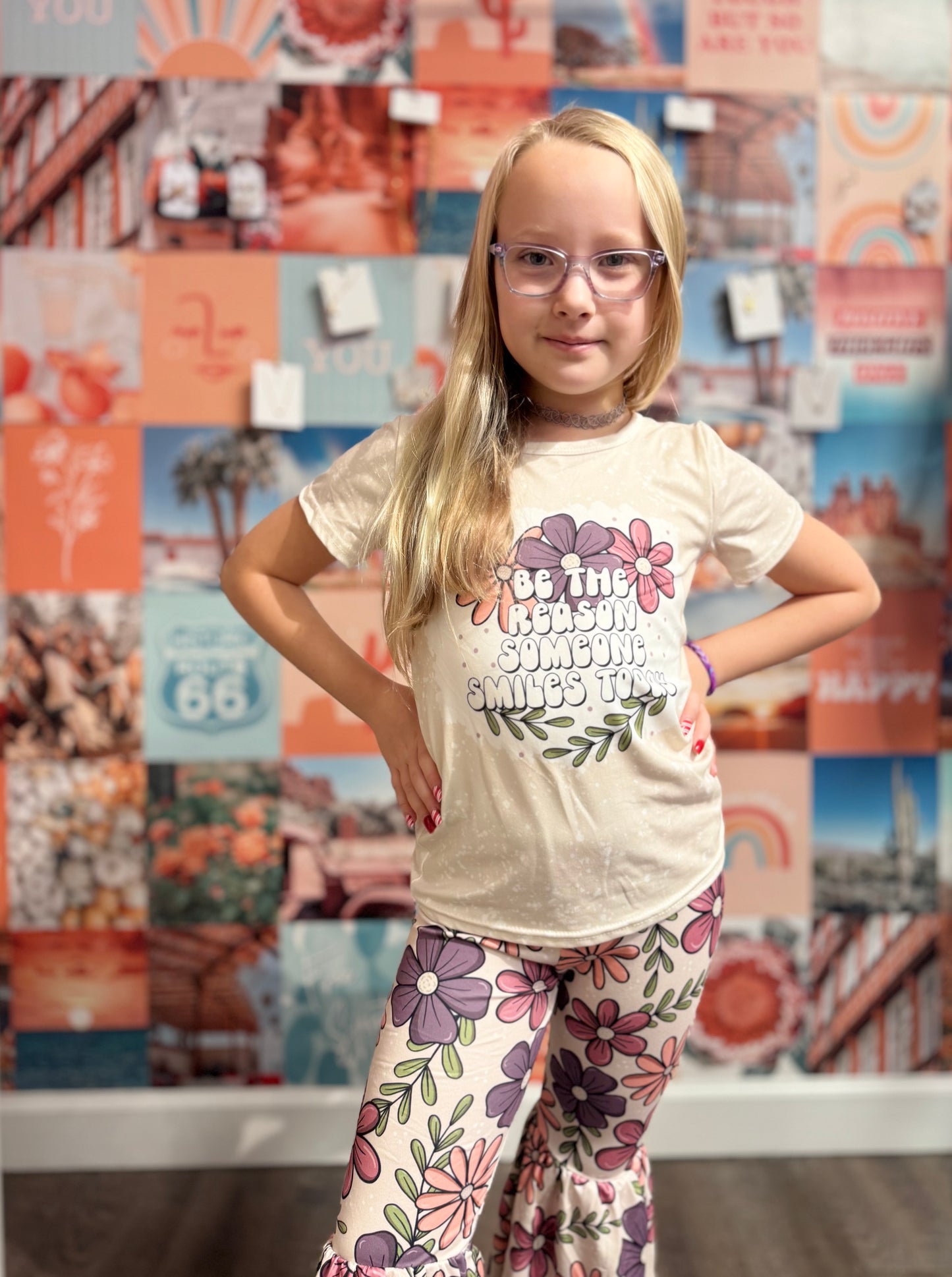 Beige short-sleeve T-shirt with a splatter design, featuring colorful floral graphics and the motivational phrase 'Be the reason someone smiles today' in bold, playful font. Paired with matching floral-print bell-bottom pants, this cheerful outfit is displayed on a pink hanger in a boutique setting, surrounded by vibrant accessories and other clothing items.