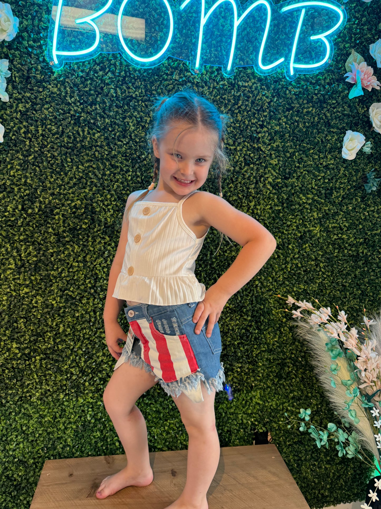 A young girl posing indoors in front of a green floral wall with a neon "BOMB" sign. She wears a white sleeveless button-up top with a ruffled hem and distressed denim shorts featuring a patriotic flag design.