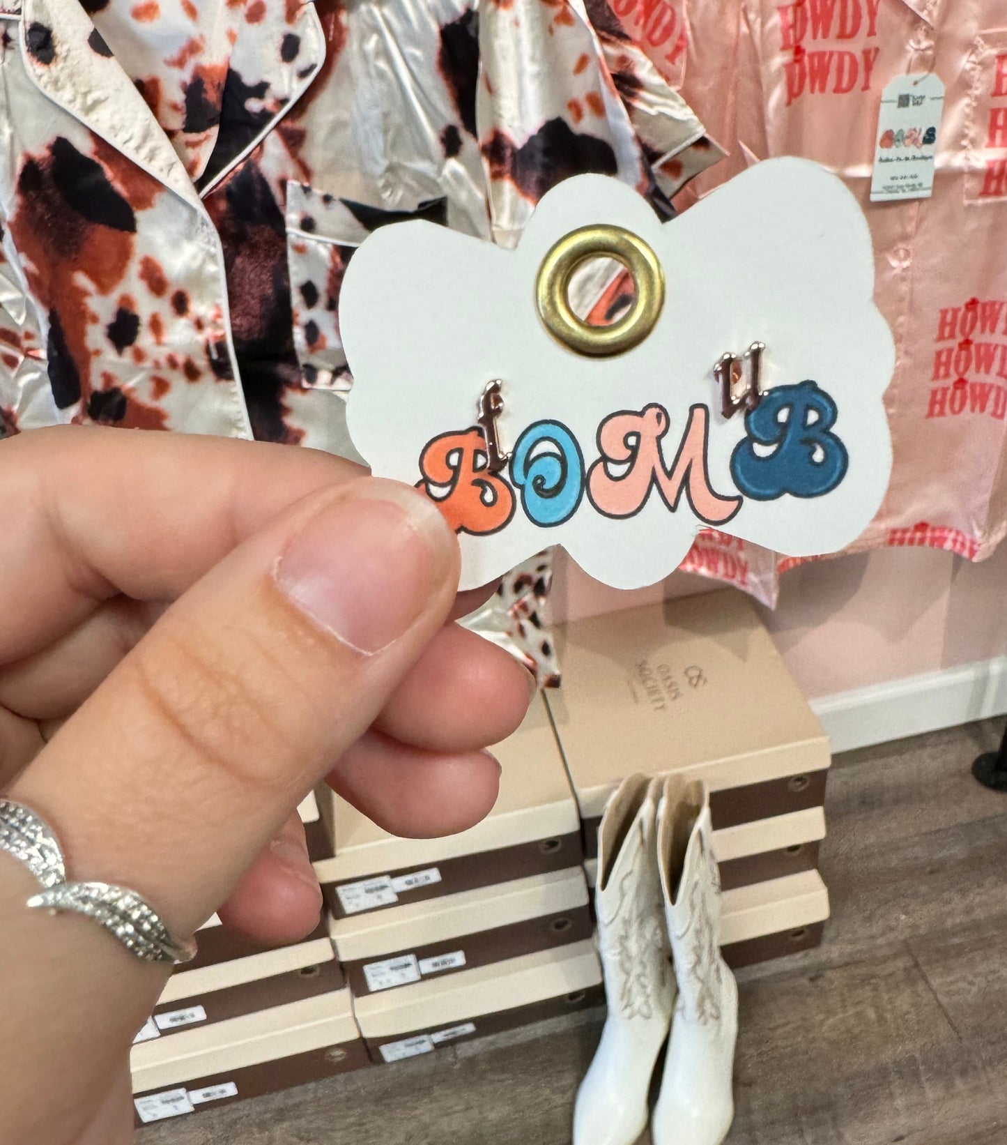 Playful earrings spelling out "F, U," displayed on a scalloped card with a gold grommet. Background features Western-themed satin apparel and white cowboy boots, adding a trendy, chic vibe.