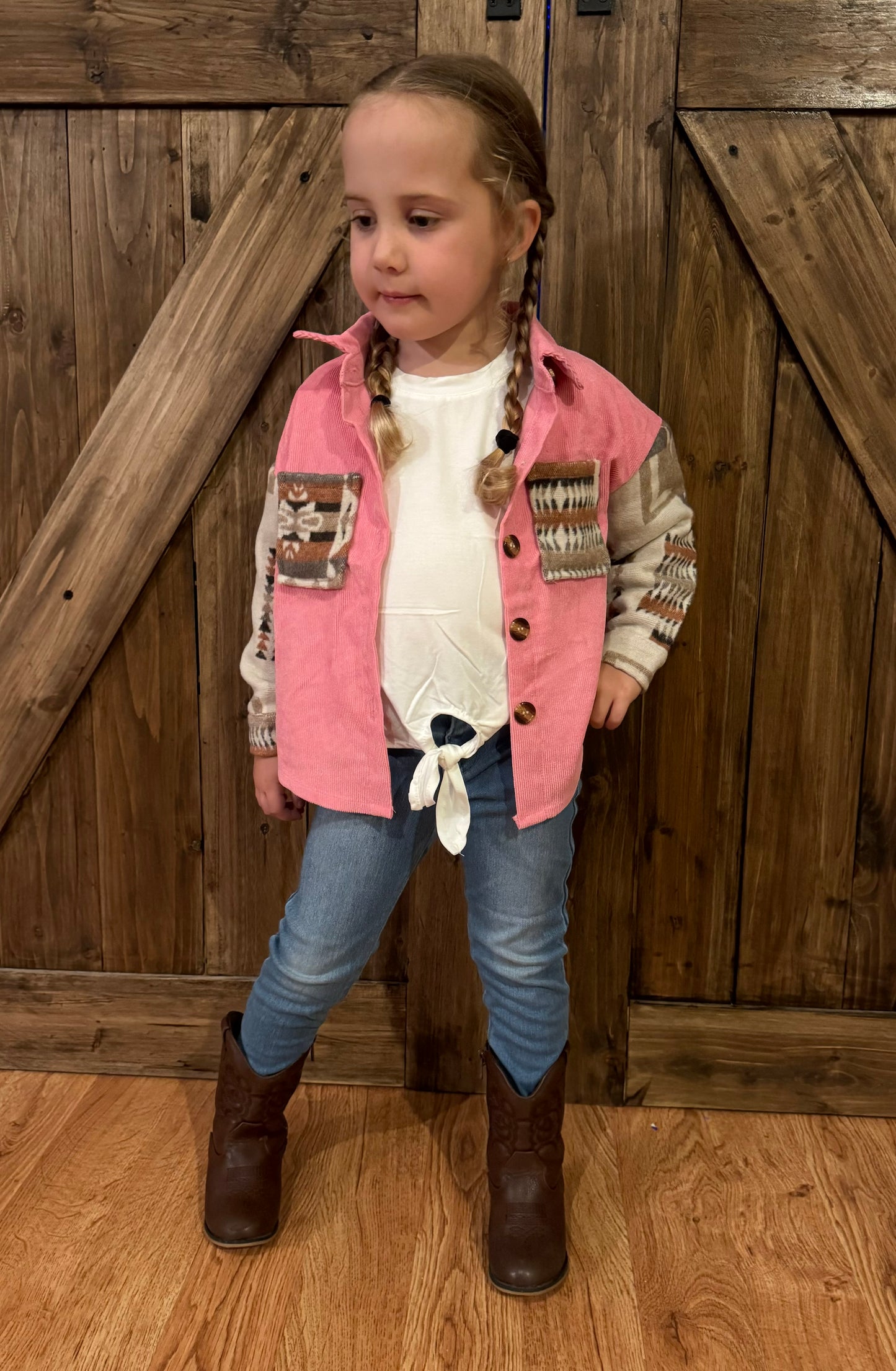 "Child wearing a pink corduroy shacket with patterned sleeves and pockets, paired with a white knotted top, blue jeans, and brown cowboy boots, standing in front of a wooden barn door."