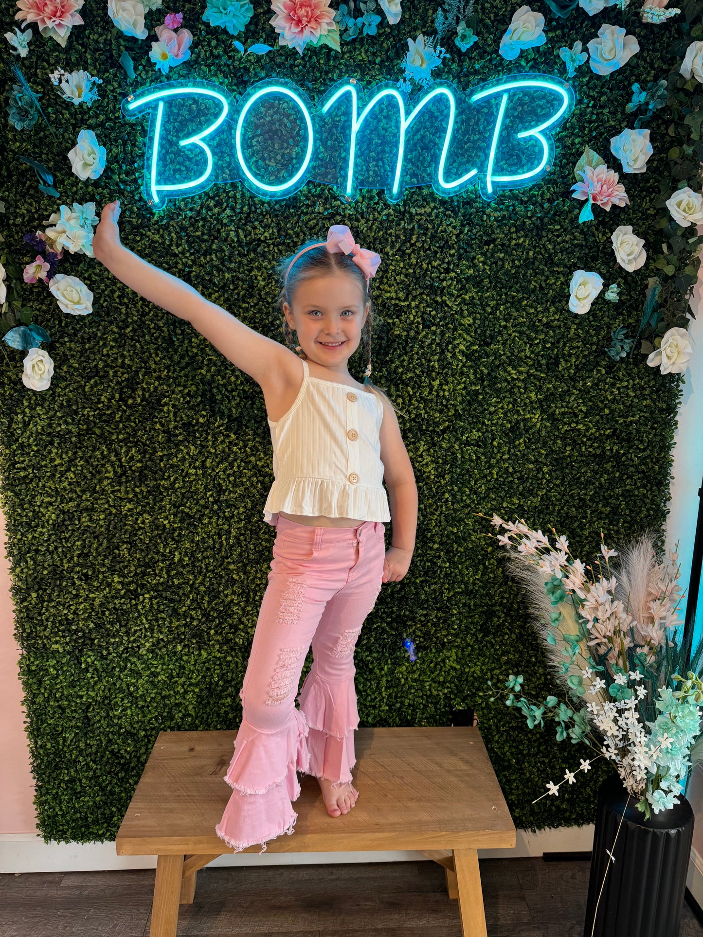 The same girl strikes a playful pose indoors, raising one arm high while standing barefoot on a wooden platform. She wears the white sleeveless top and pink bell-bottom pants and has her hair accessorized with a pink bow. She is framed by a decorative green faux foliage wall adorned with flowers, with a bright blue neon sign reading "BOMB" glowing above her. A floral arrangement in a black vase adds an elegant touch to the scene.