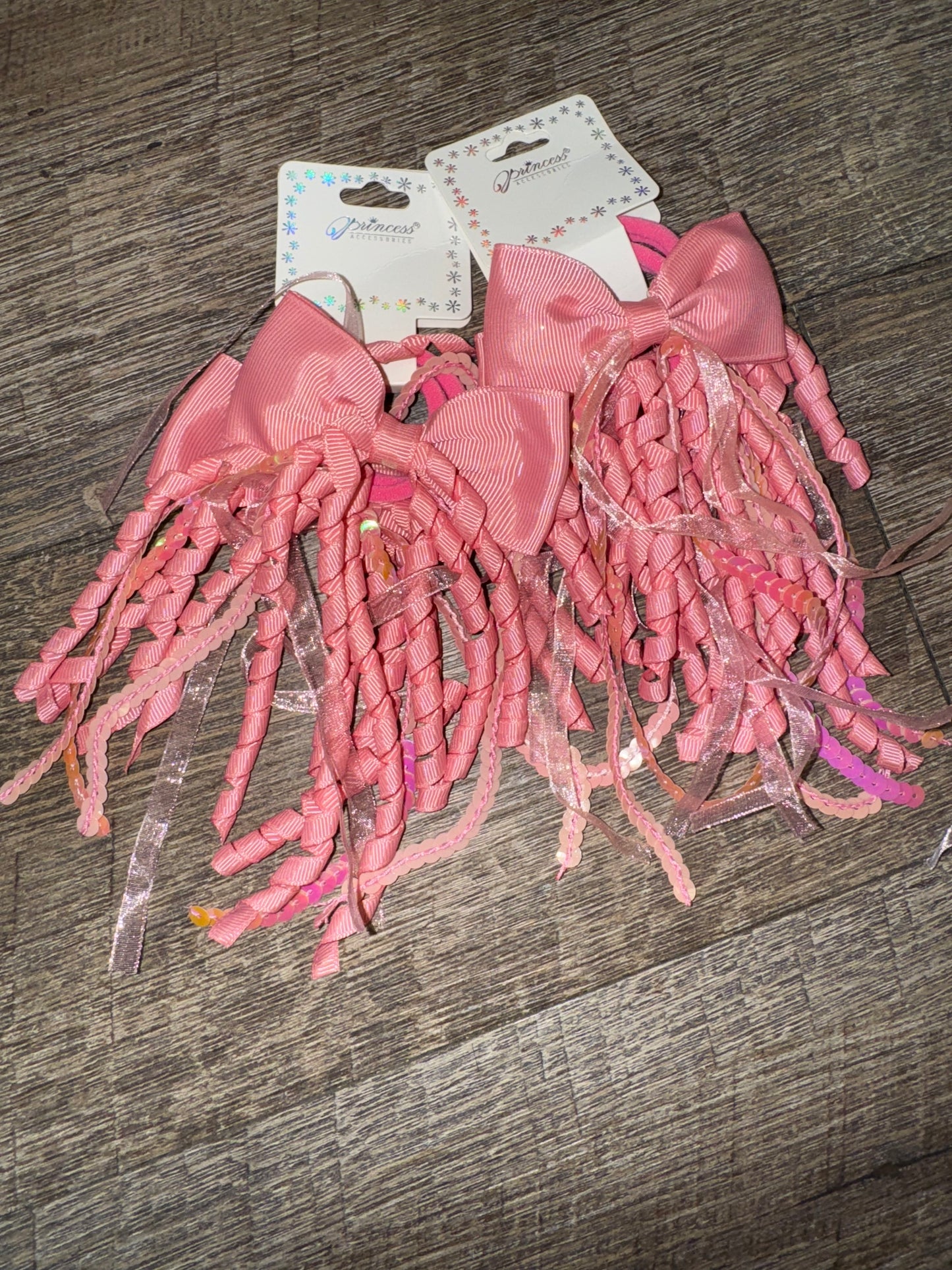 A set of light pink hair bows with cascading curly ribbons and sparkly sequin details, displayed on branded packaging labeled "Princess Accessories." The bows are placed on a wooden surface, showcasing their whimsical and elegant design.