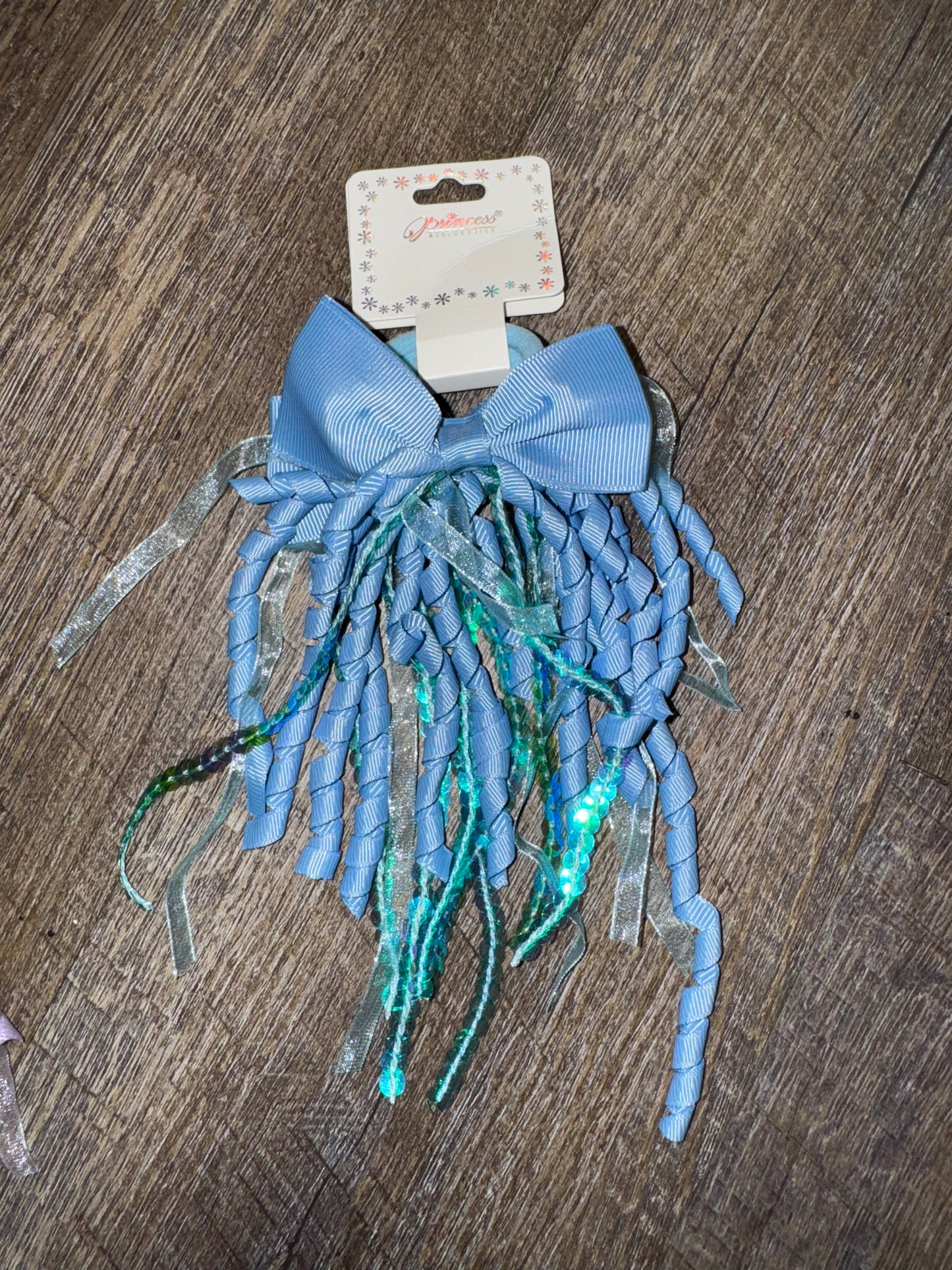 A set of blue hair bows with cascading curly ribbons and sparkly sequin details, displayed on branded packaging labeled "Princess Accessories." The bows are placed on a wooden surface, showcasing their whimsical and elegant design.