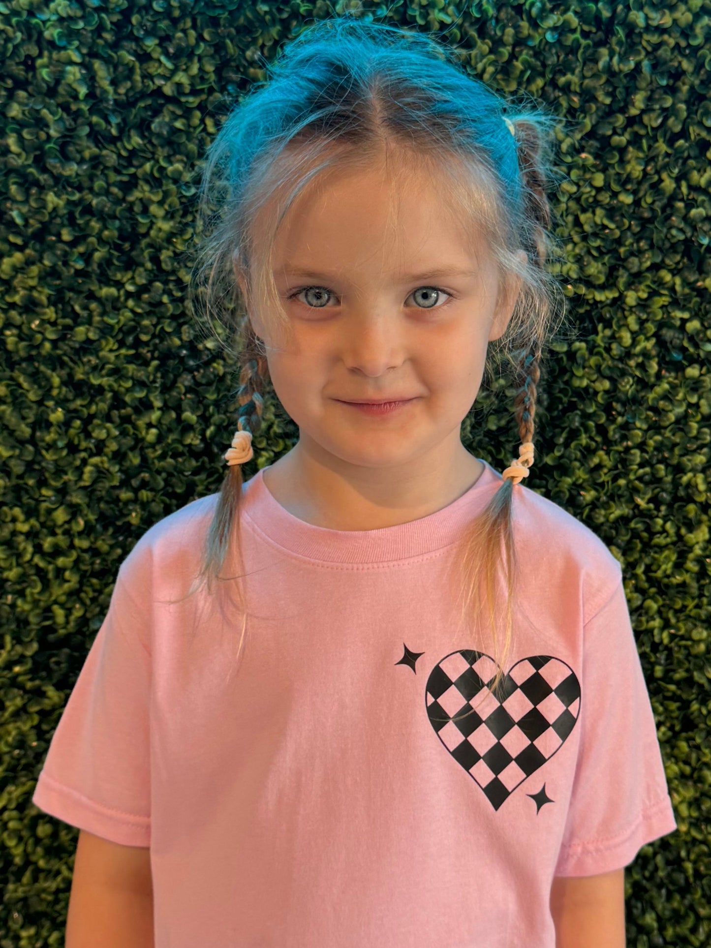 A young girl with light blonde hair styled in two braided pigtails, wearing a light pink t-shirt featuring a black and white checkered heart graphic with small sparkling accents. She is standing in front of a vibrant green leafy wall, which creates a natural textured background. The soft lighting highlights her bright blue eyes and faint smile, creating a warm and cheerful atmosphere.
