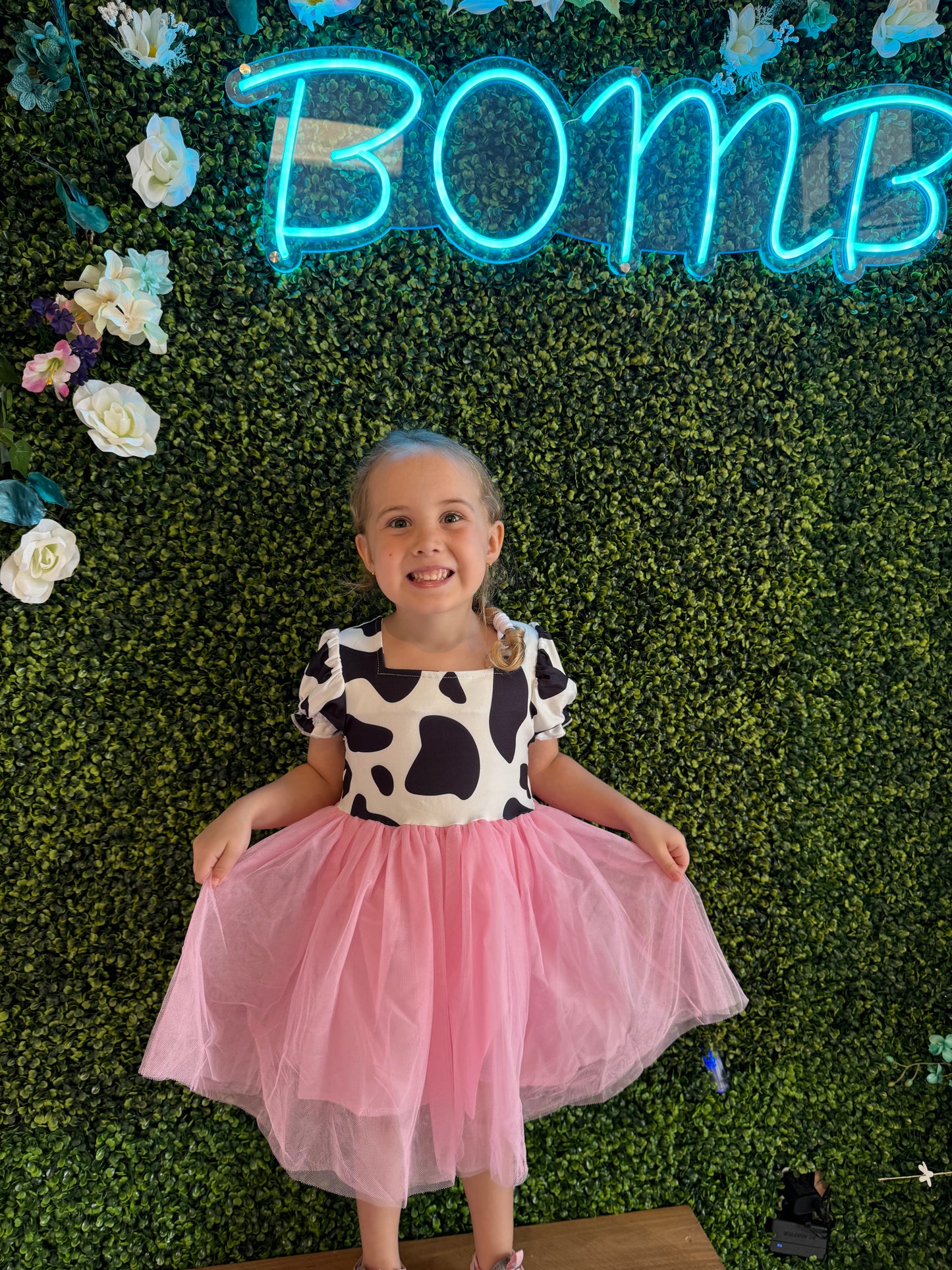 A young girl smiles brightly while wearing a playful dress with a cow-print bodice featuring black spots on a white background and puffed sleeves. The dress has a flowing pink tulle skirt that adds a whimsical touch. She stands barefoot on a wooden platform in front of a green faux foliage wall adorned with white and pastel flowers. Above her, a glowing neon blue sign reads "BOMB," adding a trendy, fun vibe to the setting.