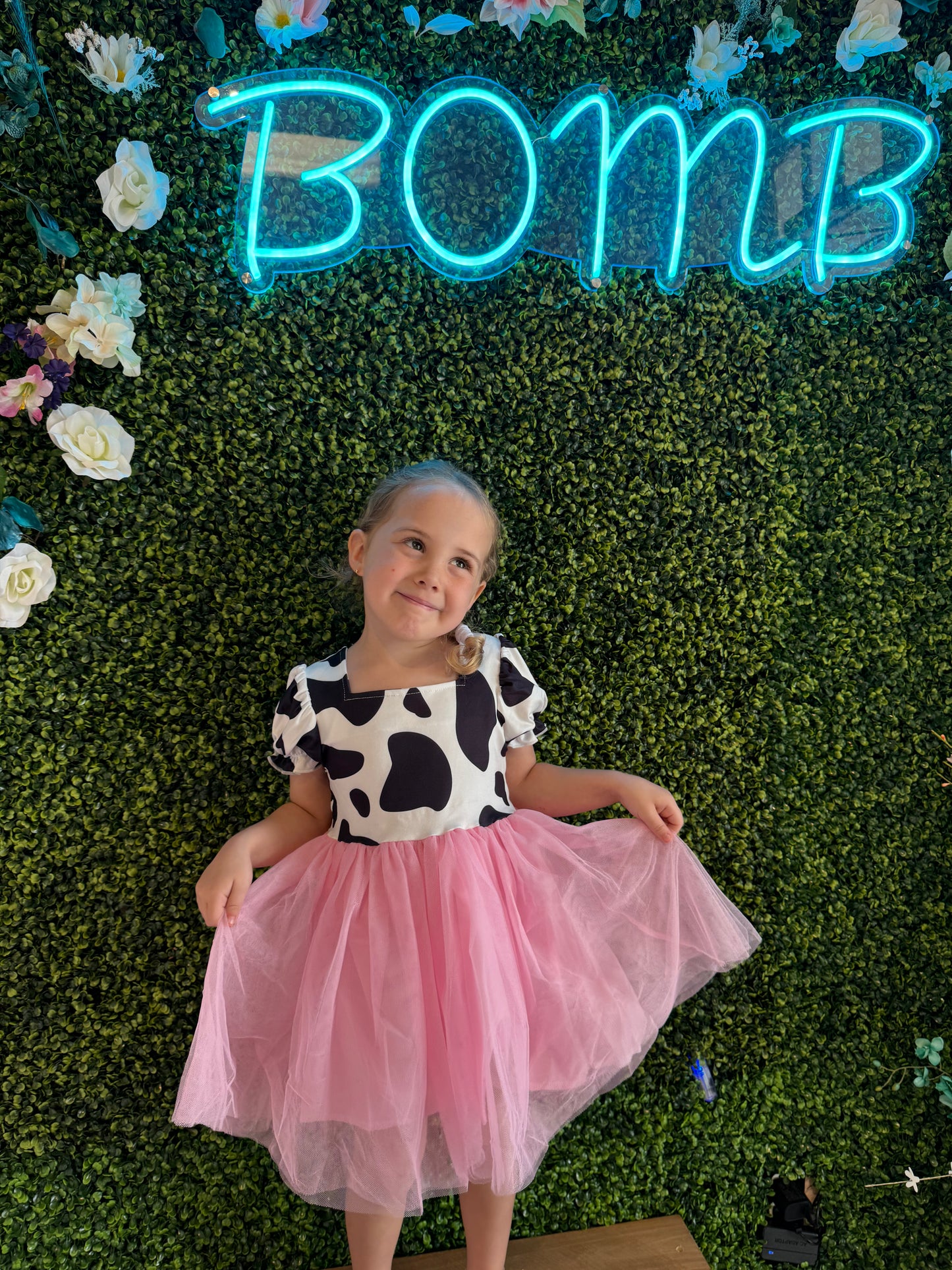 A young girl poses sweetly, tilting her head with a playful smile while wearing a dress with a cow-print bodice featuring black spots on a white background and puffed sleeves. The outfit is completed with a voluminous pink tulle skirt, creating a charming and whimsical look. She stands barefoot on a wooden platform, set against a lush green faux foliage wall decorated with white and pastel flowers. Above her, a vibrant blue neon sign reads "BOMB," adding a modern and lively touch to the scene.