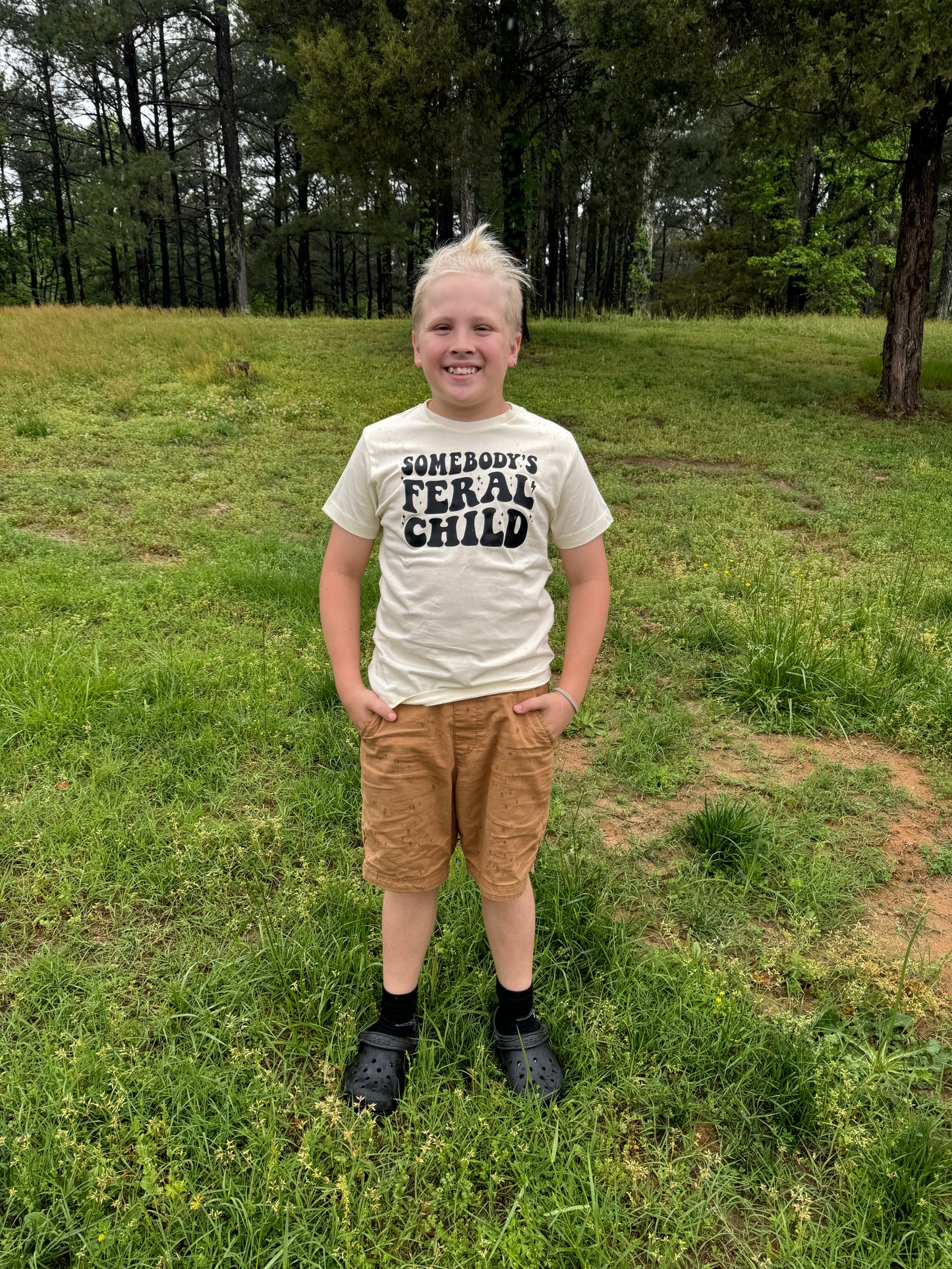 A close-up view of a child wearing an off-white t-shirt featuring bold black text that reads "Somebody's Feral Child." The child stands outdoors on a grassy lawn, their hands tucked into tan pants.