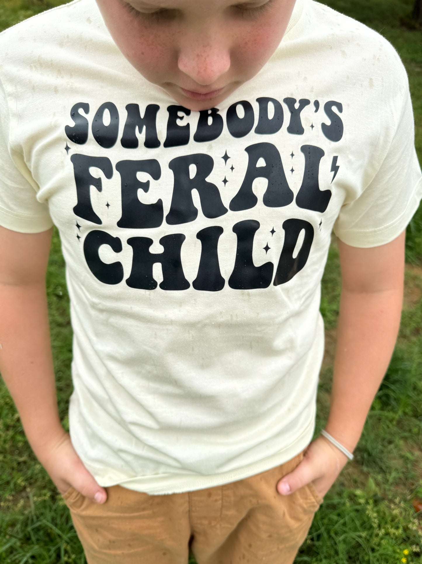 A close-up view of a child wearing an off-white t-shirt featuring bold black text that reads "Somebody's Feral Child." The child stands outdoors on a grassy lawn, their hands tucked into tan pants.