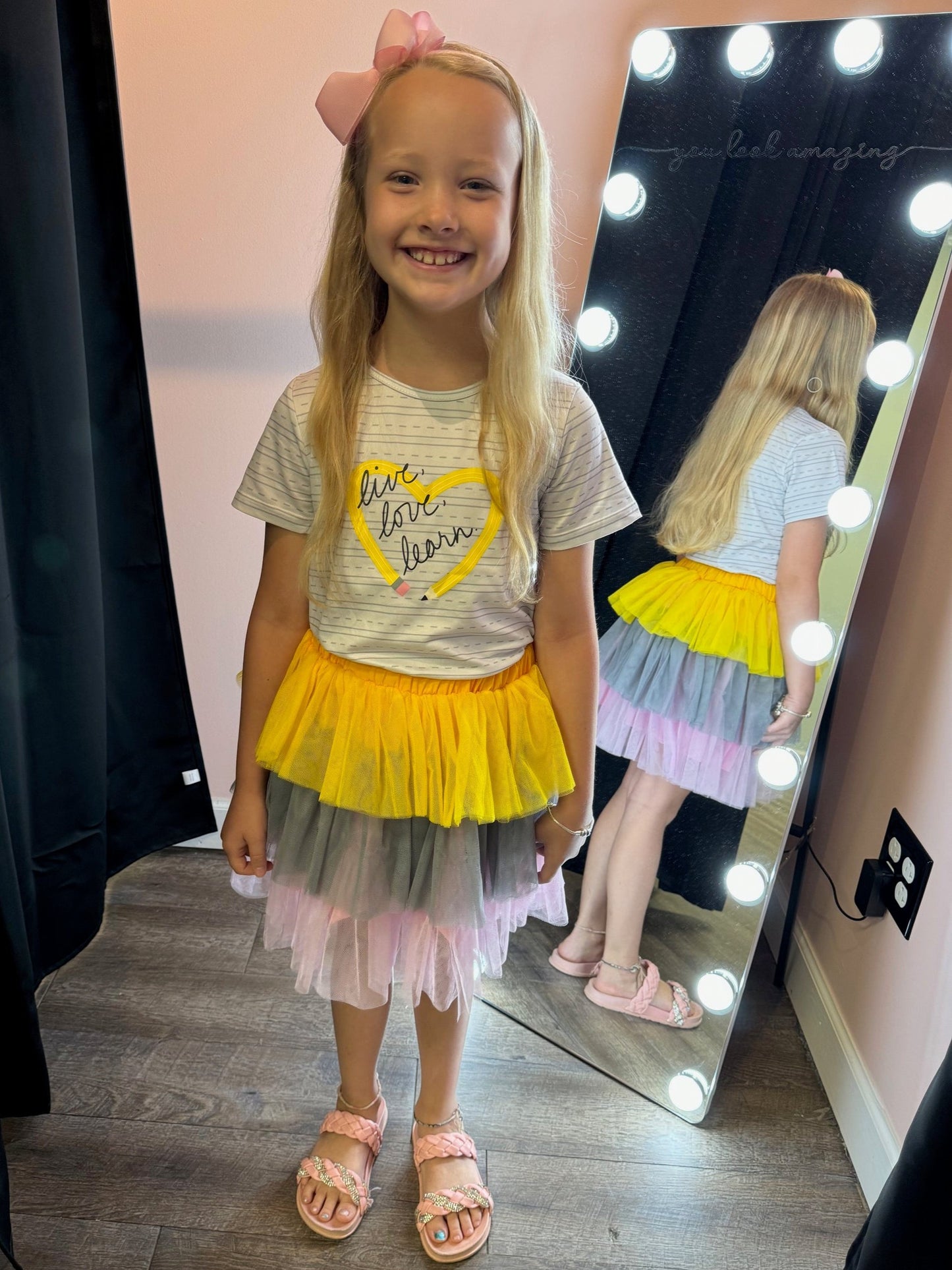 Smiling young girl standing in front of a vanity mirror with round lights, wearing a white T-shirt with the phrase 'live, love, learn' inside a yellow heart and a layered tulle skirt in yellow, gray, and pink tones. She has long blonde hair adorned with a pink bow, paired with pink braided sandals. The background features a wooden floor and a softly lit wall, creating a cheerful and stylish atmosphere.