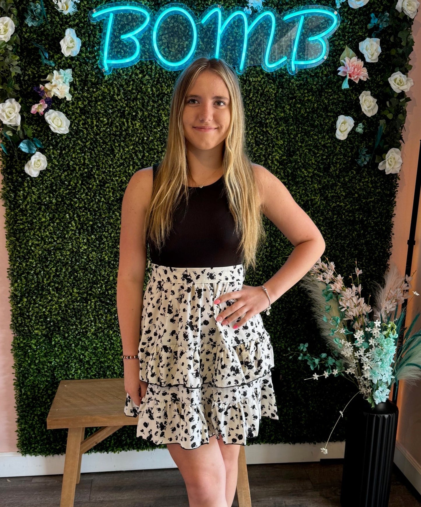 A young woman poses confidently in front of a decorative backdrop featuring a green faux boxwood wall adorned with white and pink flowers, with a glowing neon "BOMB" sign at the top. She wears a sleeveless black top tucked into a white tiered skirt with a black floral pattern, creating a chic and feminine look. Her long, straight blonde hair is styled naturally, and her accessories include a delicate bracelet on her wrist. A wooden stool and a black vase with pastel floral accents are visible in the stylish