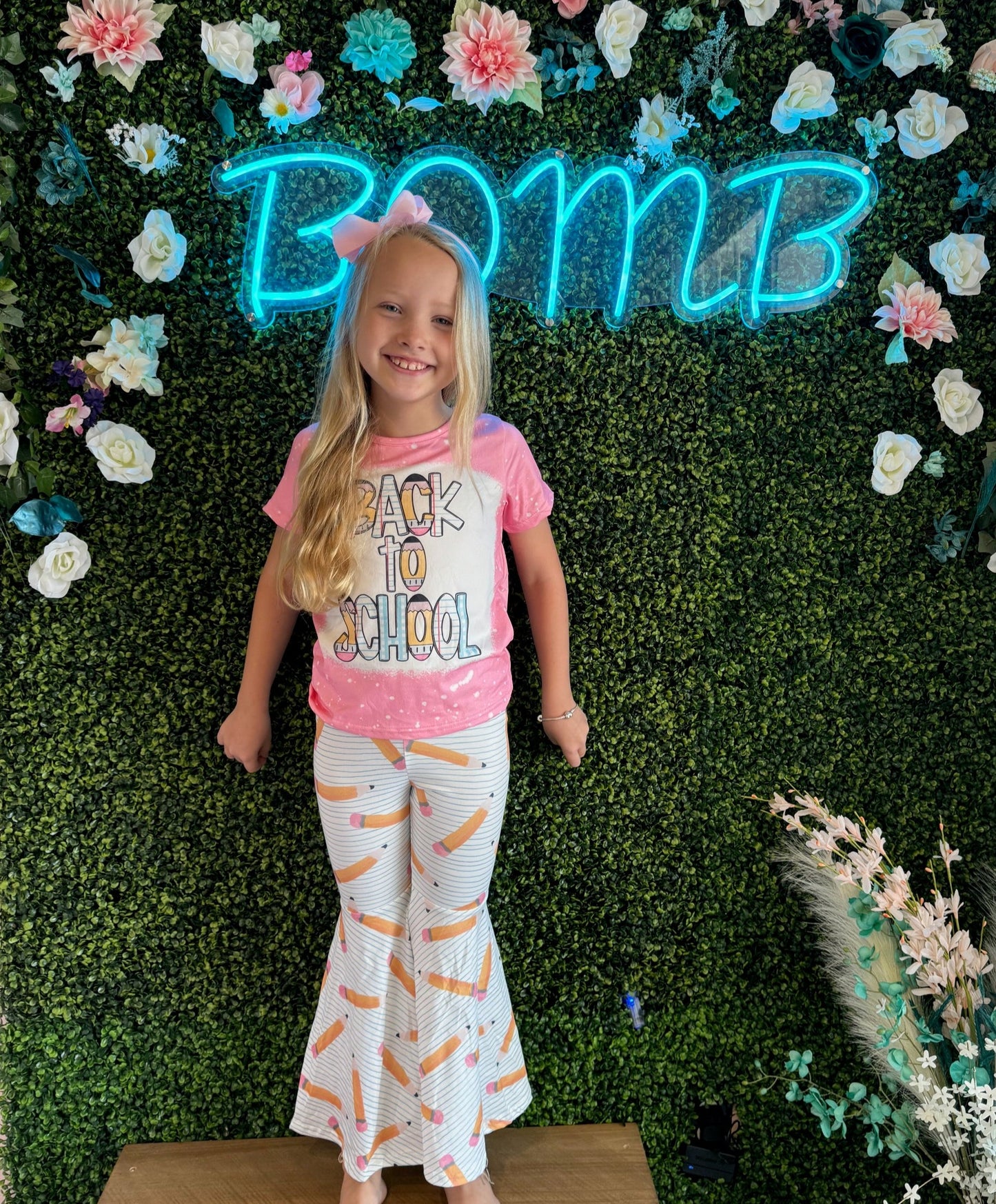 Smiling young girl in a pink 'Back to School' T-shirt and white bell-bottom pants featuring a pencil print, standing against a lush green faux foliage wall decorated with flowers and a glowing blue 'BOMB' neon sign. She completes her look with a pink hair bow, radiating back-to-school excitement.