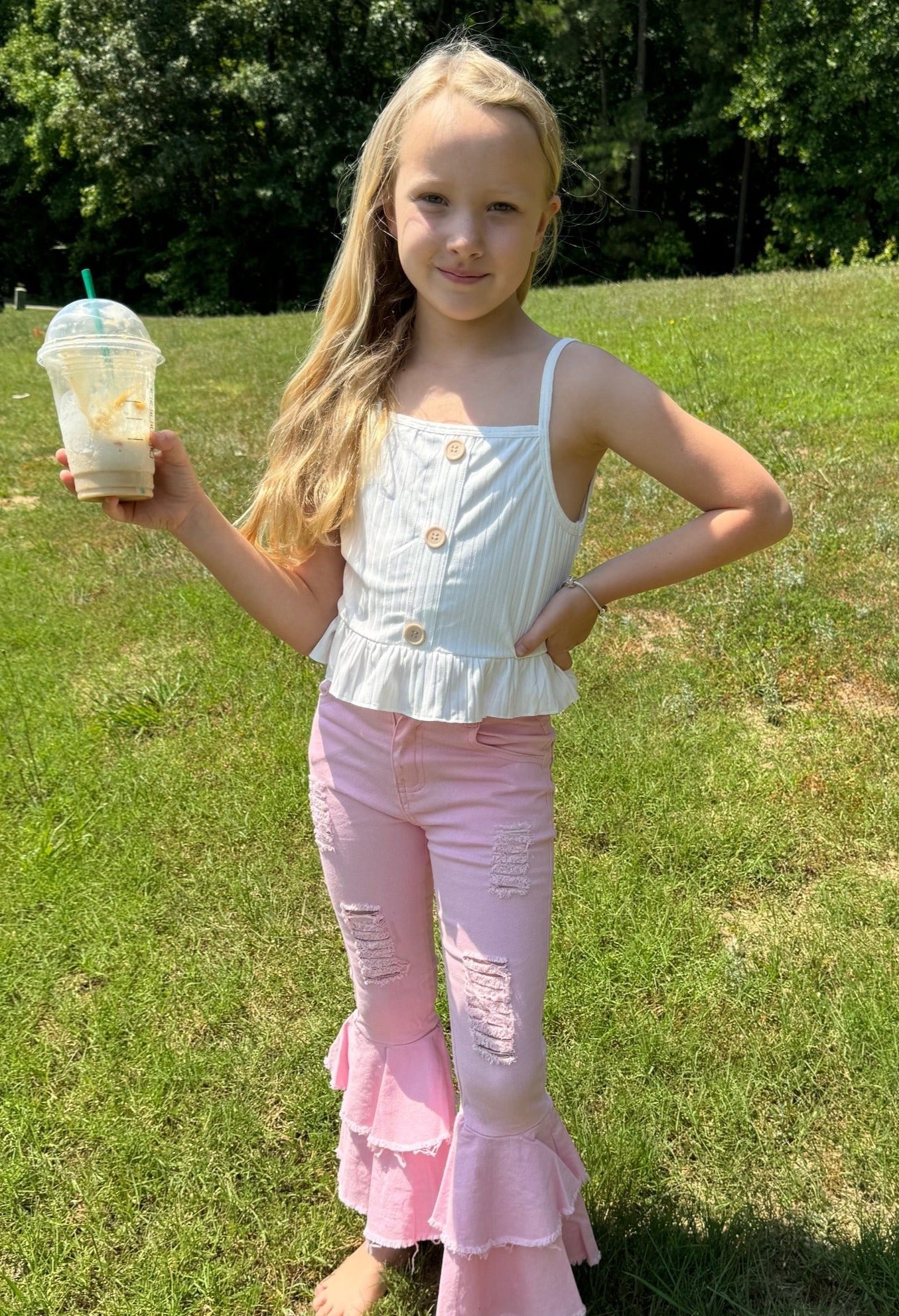A young girl stands confidently outdoors, holding a partially finished iced drink in a clear cup with a green straw. She wears a sleeveless white top with wooden buttons and a ruffled hem, paired with light pink bell-bottom pants featuring distressed patches and tiered ruffles at the bottom. She is barefoot on a lush green lawn, enjoying the sunny day.