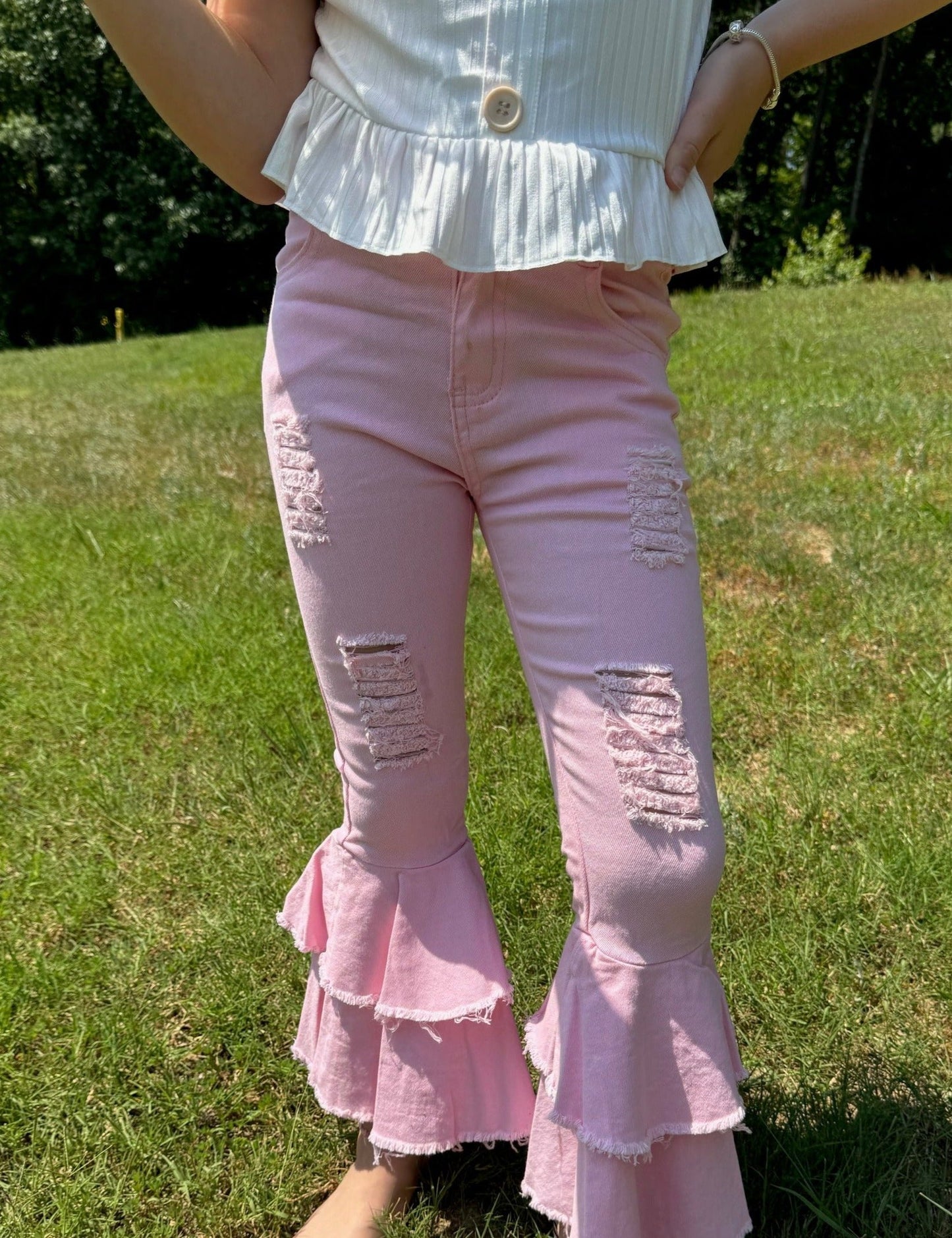 A close-up view of the girl's light pink bell-bottom pants, showcasing their distressed details on the thighs and the layered, frayed ruffles at the bottom. The pants are paired with a white ruffled top, with the background showing a grassy outdoor setting.