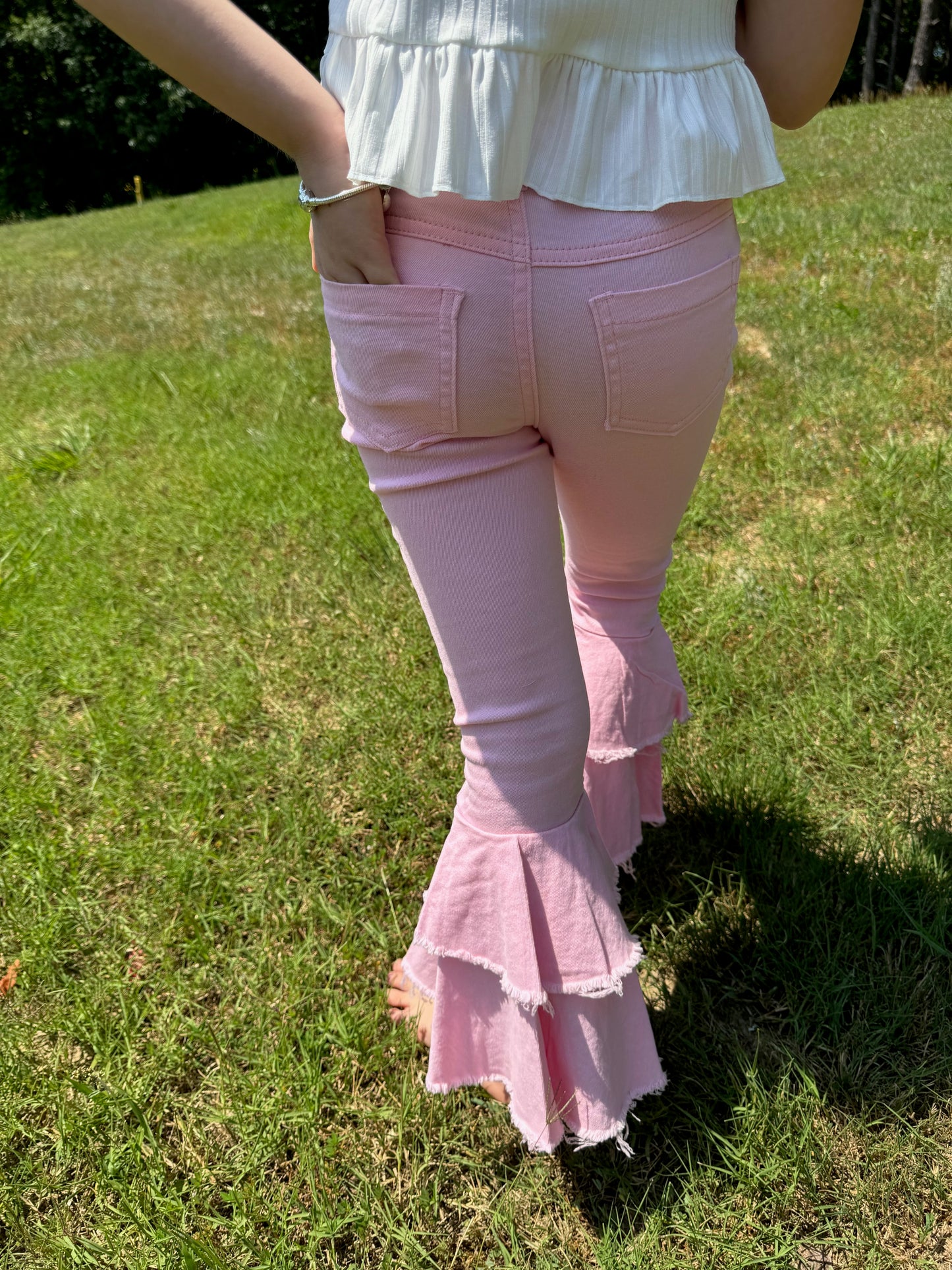 A rear view of a young girl wearing light pink bell-bottom jeans with frayed tiered ruffles at the ankles. The jeans feature functional back pockets and a fitted design that highlights their trendy, boho-inspired style. She pairs the pants with a white ruffled top and stands barefoot on lush green grass, with sunlight casting soft shadows around her. The outfit exudes a playful and summery vibe.