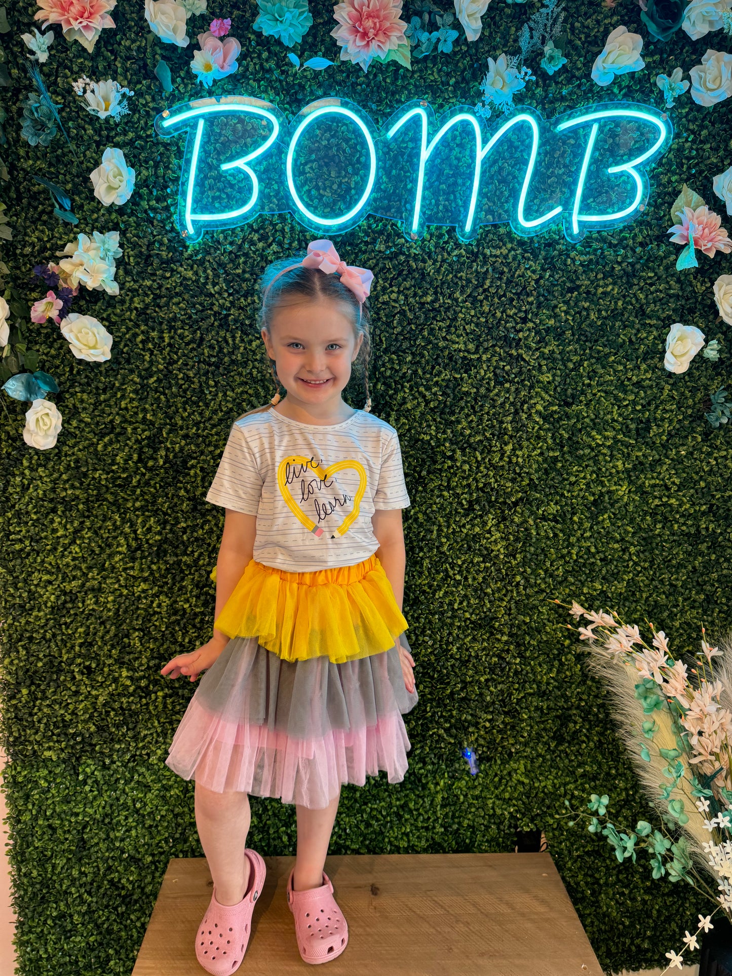A young girl standing in front of a vibrant greenery wall adorned with floral decorations and a glowing neon 'BOMB' sign, wearing a colorful tulle skirt in yellow, pink, and gray, a striped graphic tee with 'Live, Love, Learn,' and pink Crocs. The stylish boutique background reflects trendy and family-friendly fashion vibes.
