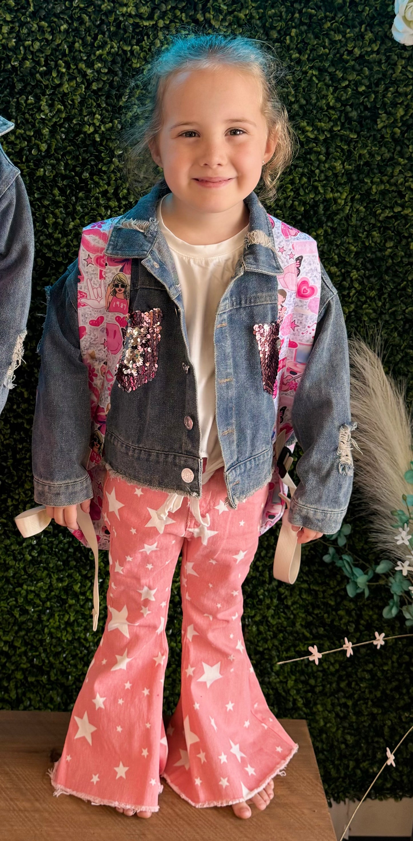 A young girl smiles confidently, wearing a stylish outfit that includes a distressed denim jacket with sparkling pink sequin patches on the chest. The jacket is paired with pink bell-bottom pants adorned with a bold white star pattern and frayed hems. Underneath the jacket, she wears a simple white top, and she carries a pink-themed backpack with playful graphics featuring hearts, sunglasses, and fun designs. She stands barefoot on a wooden surface in front of a lush green faux foliage wall, creating a whim