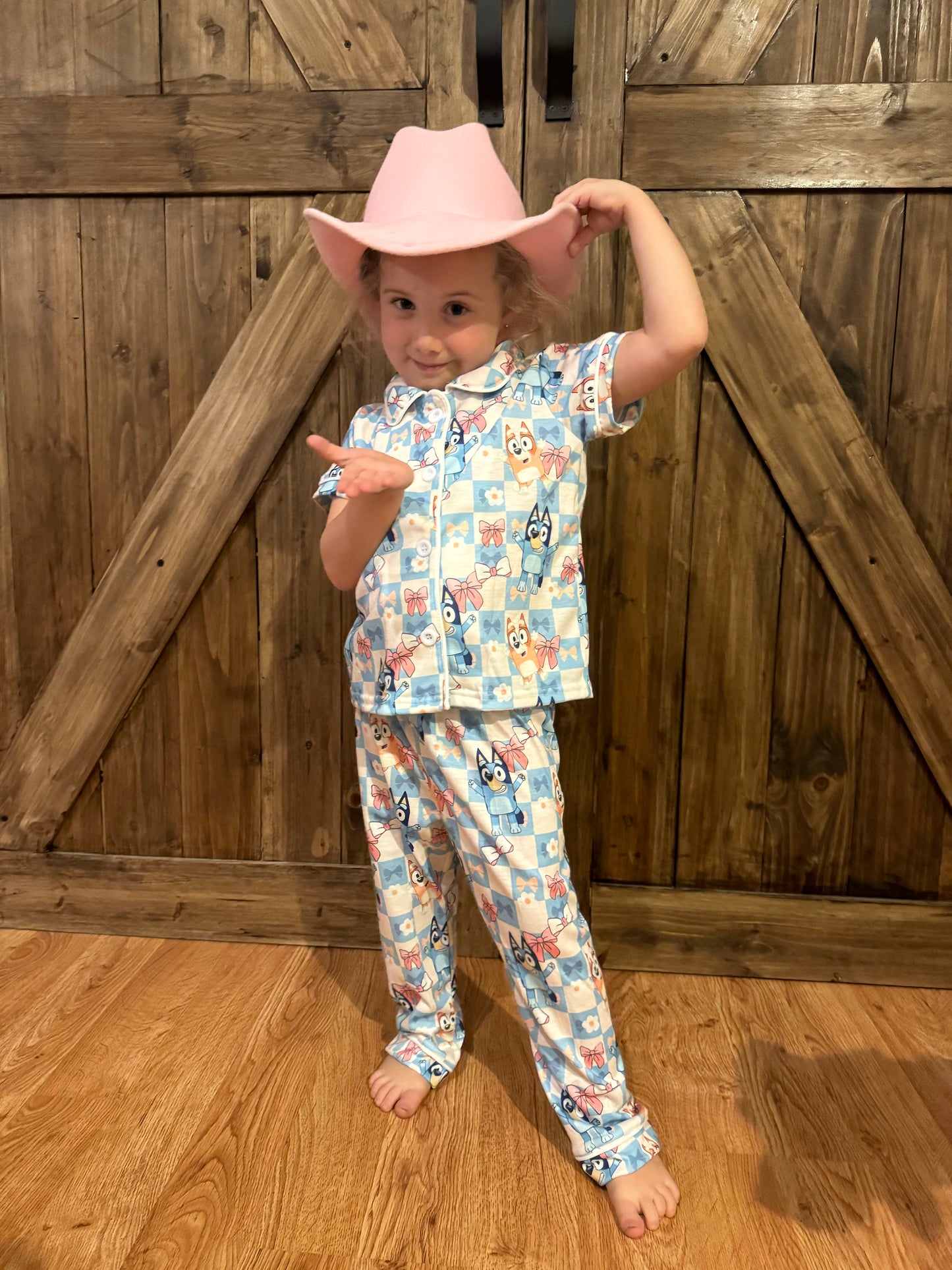 The same young girl wears light blue pajama sets adorned with bluey and bingo, bows, and flowers in a pastel checkered pattern. She pairs the outfit with a pink cowboy hat, posing with one arm raised to touch the hat and the other gesturing playfully. She stands barefoot on the wood floor, framed by the same rustic wooden barn doors, adding charm and personality to the scene.