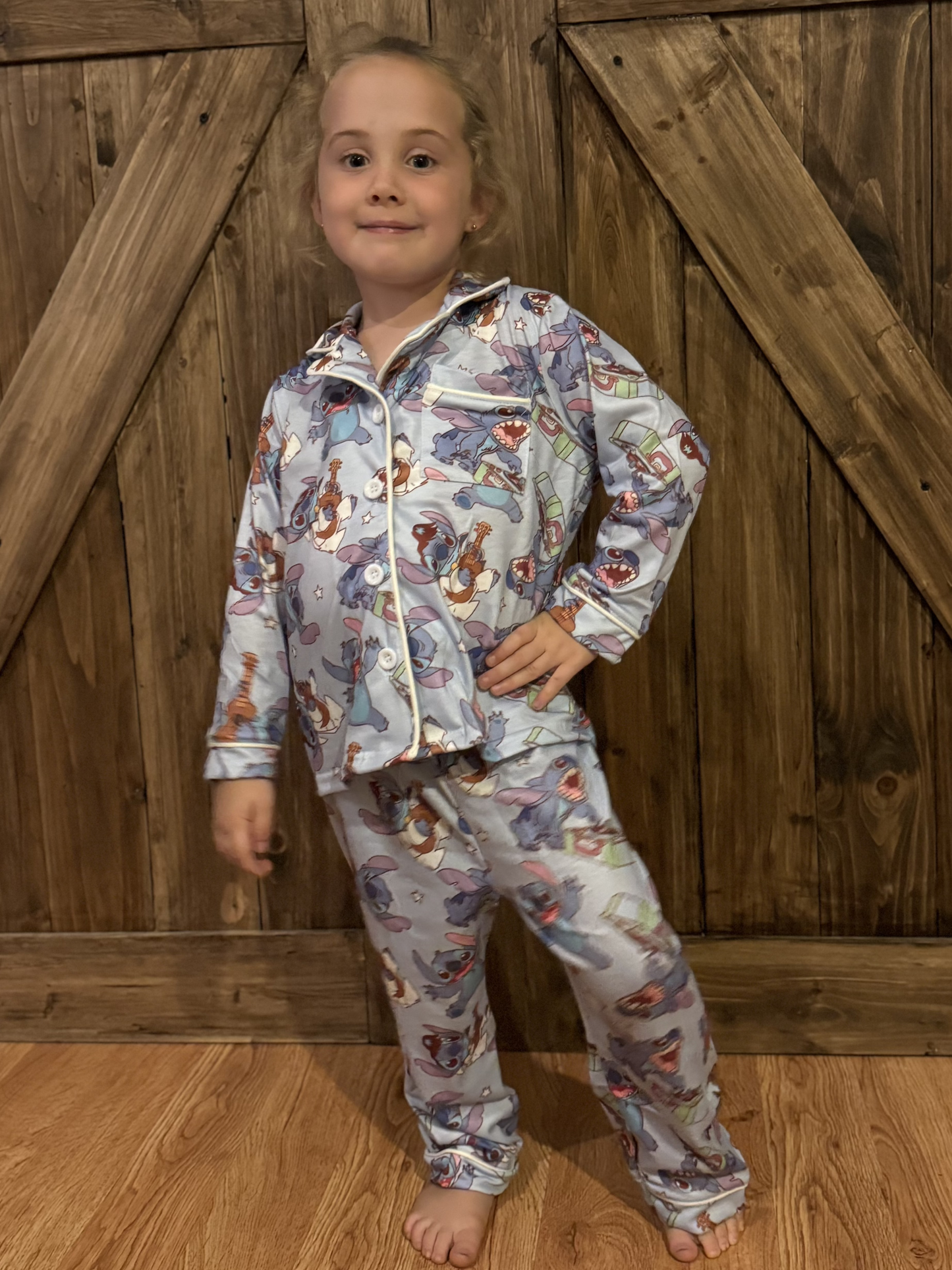 A young girl poses confidently in a cozy set of light blue pajamas featuring stitch in various whimsical poses. The long-sleeve button-up top and matching pants showcase vibrant colors and fun imagery, creating a cheerful bedtime look. She stands barefoot on a wooden floor, with rustic barn-style wooden doors as the background, giving the scene a warm and homey feel.